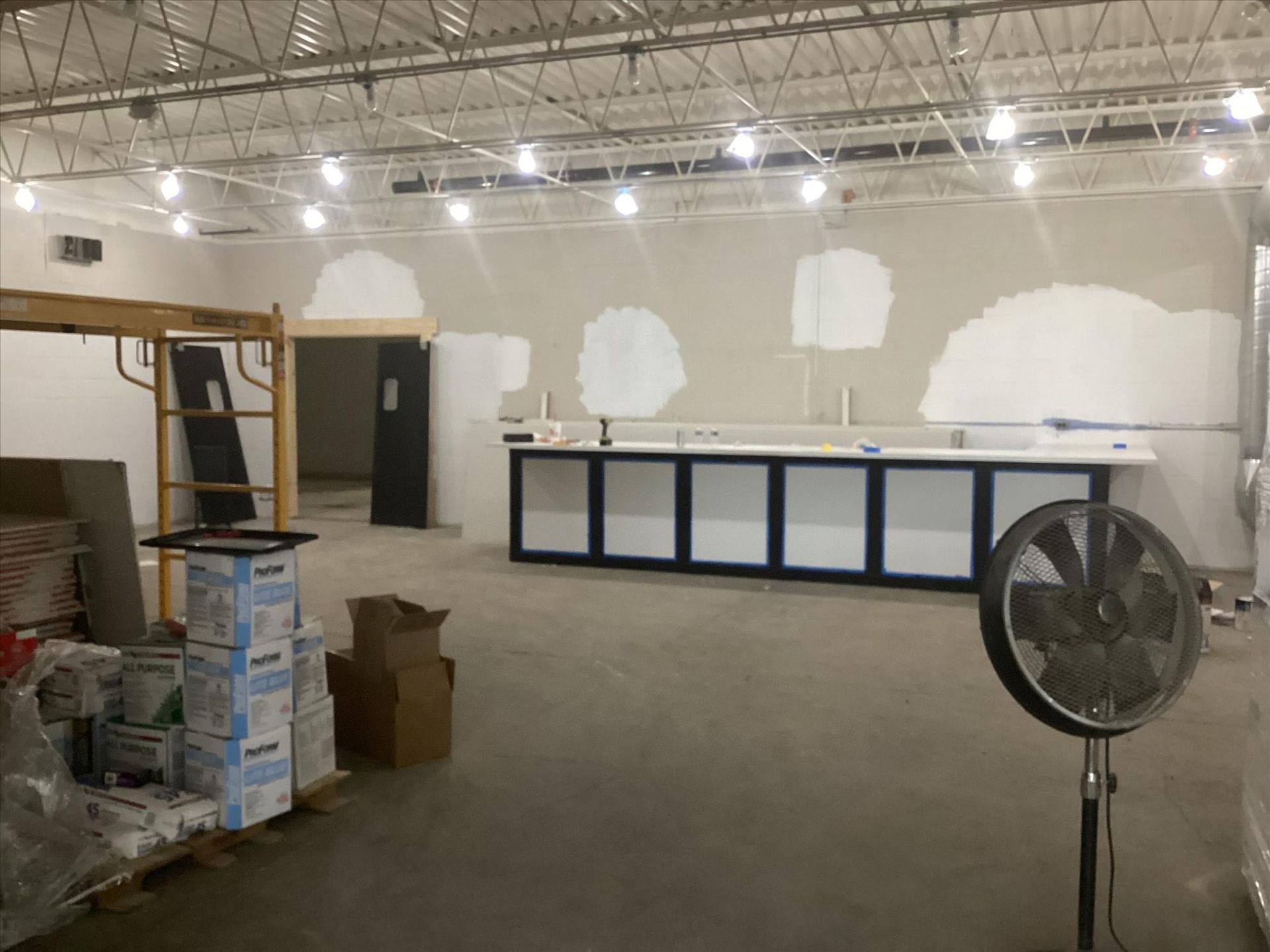 Empty commercial room with unfinished walls, stacked supplies, and a large floor fan in the foreground