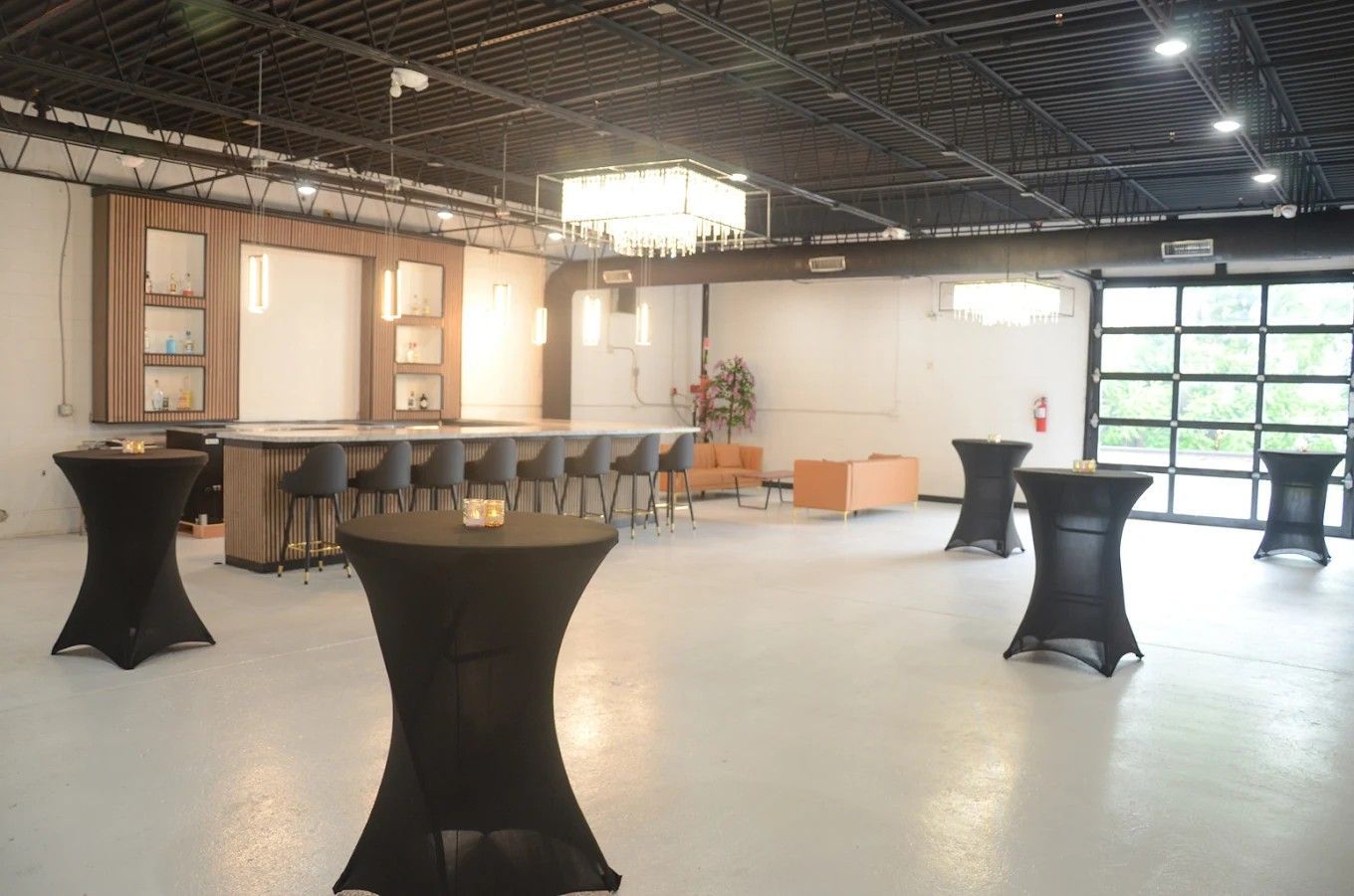 Empty event space with high tables, bar counter, exposed ceiling, and large garage door windows