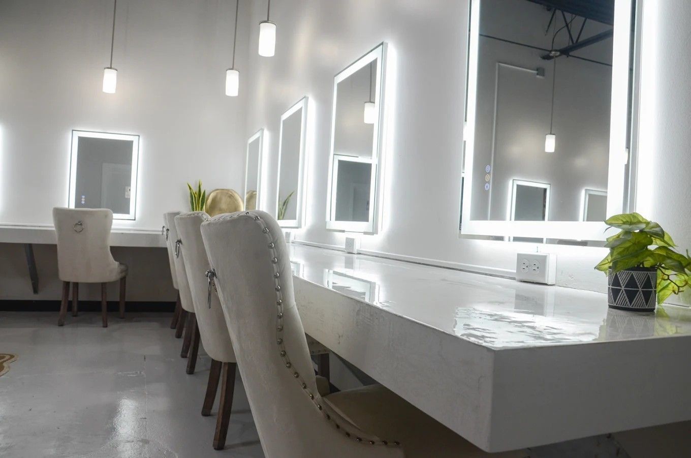 Bright modern vanity room with white desks, chairs, mirrors, and hanging pendant lights.