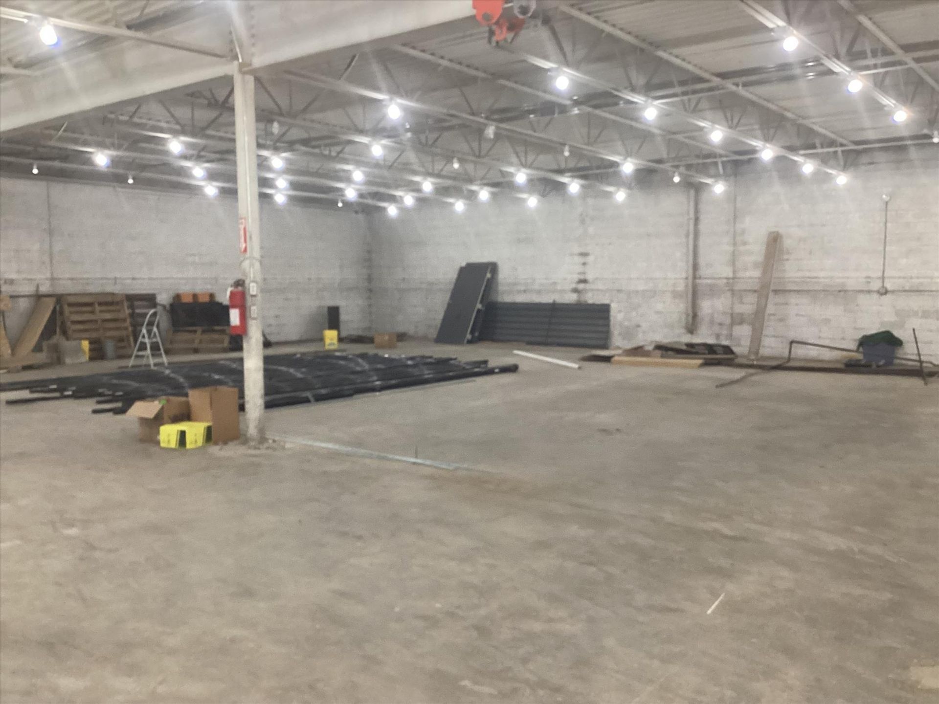 Empty warehouse interior with concrete floor, bright ceiling lights, and stacked dark materials.
