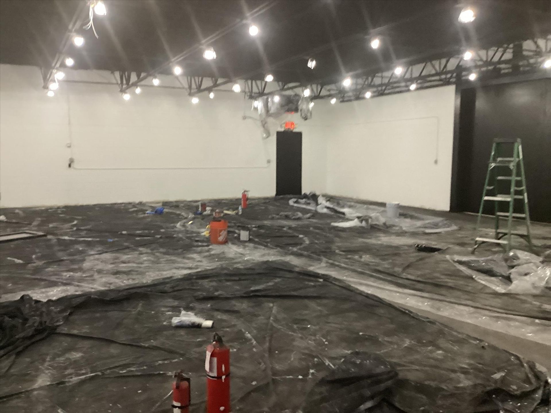 Large empty room under renovation with plastic sheeting on the floor, ladders, and bright ceiling lights