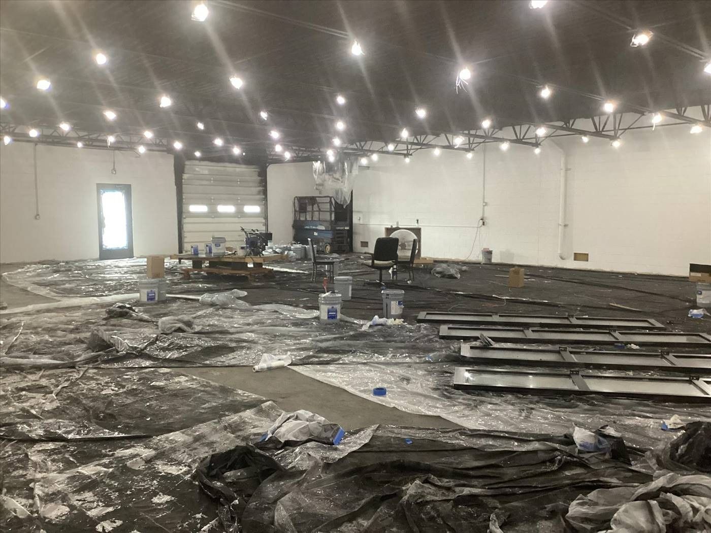 Large indoor hall under construction, with black floor covering, scattered materials, and bright ceiling lights.