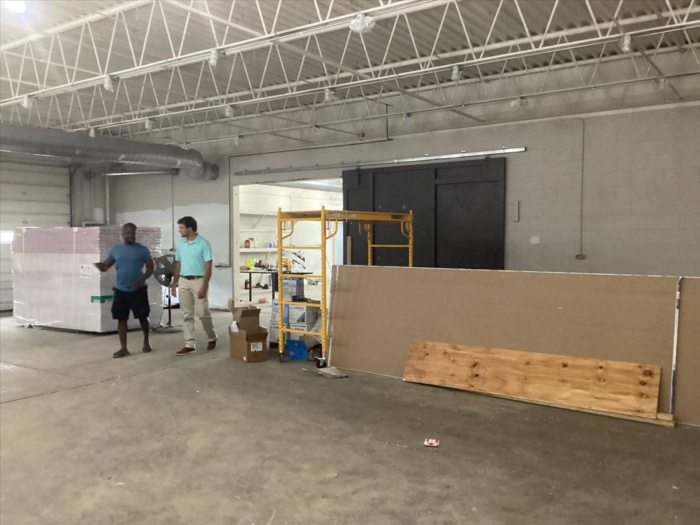 Two workers stand in a warehouse beside a large partition wall and a scaffold cart.
