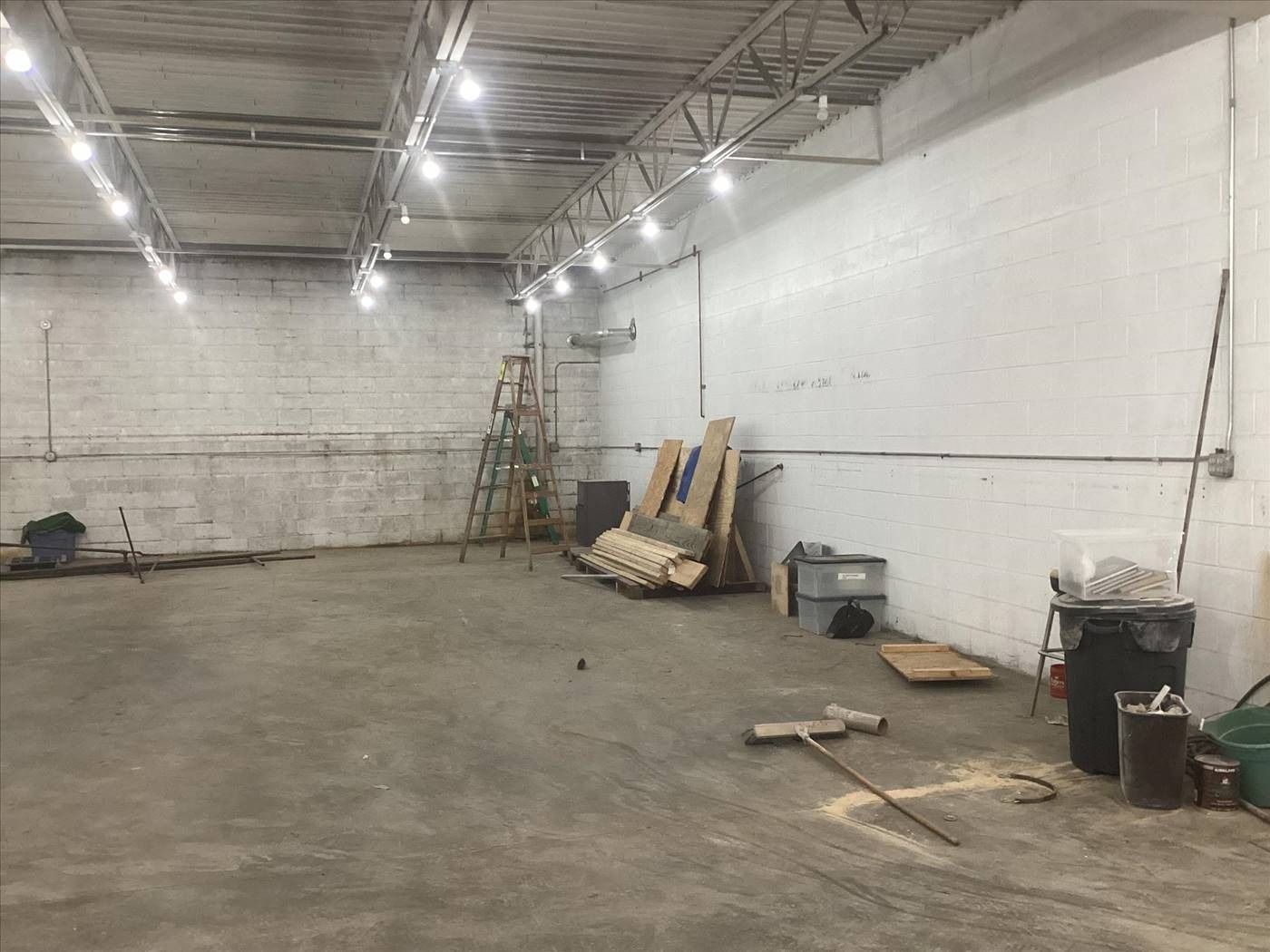 Empty warehouse interior with concrete floor, exposed ceiling lights, and construction materials stacked along the wall