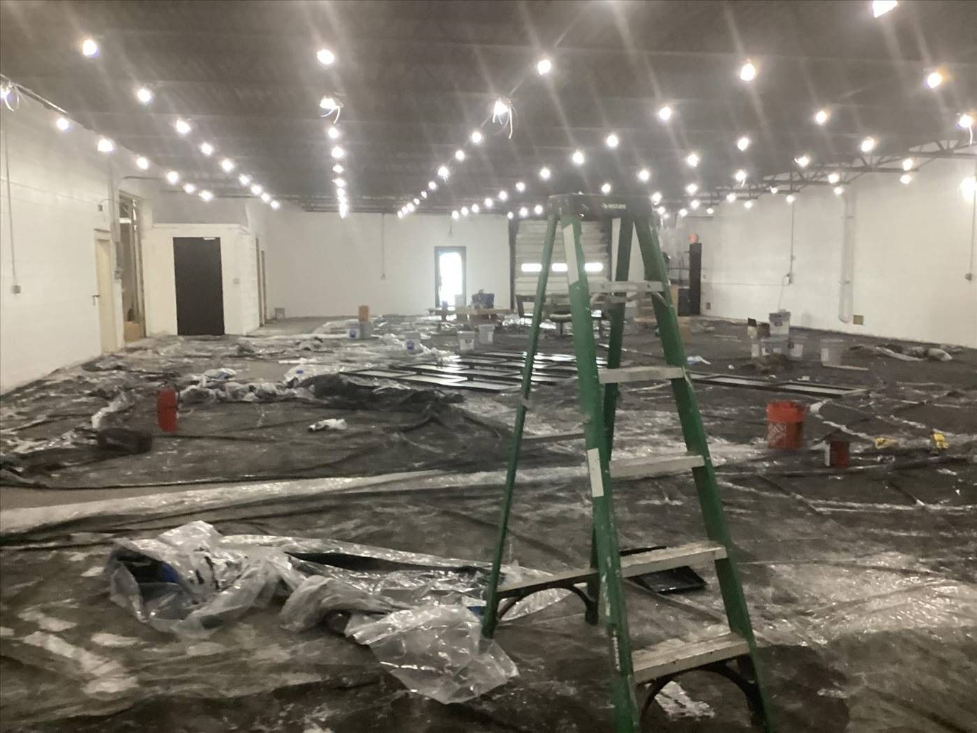 Empty indoor warehouse with debris-covered floor and a green ladder under bright ceiling lights