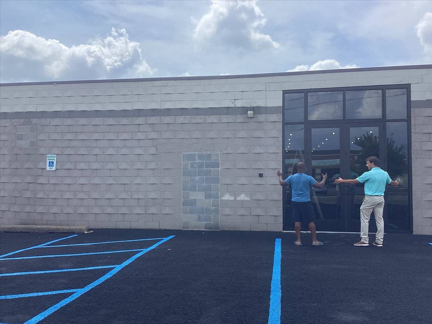 Two people at a storefront entrance on a cloudy day, with a mostly empty parking lot in front.