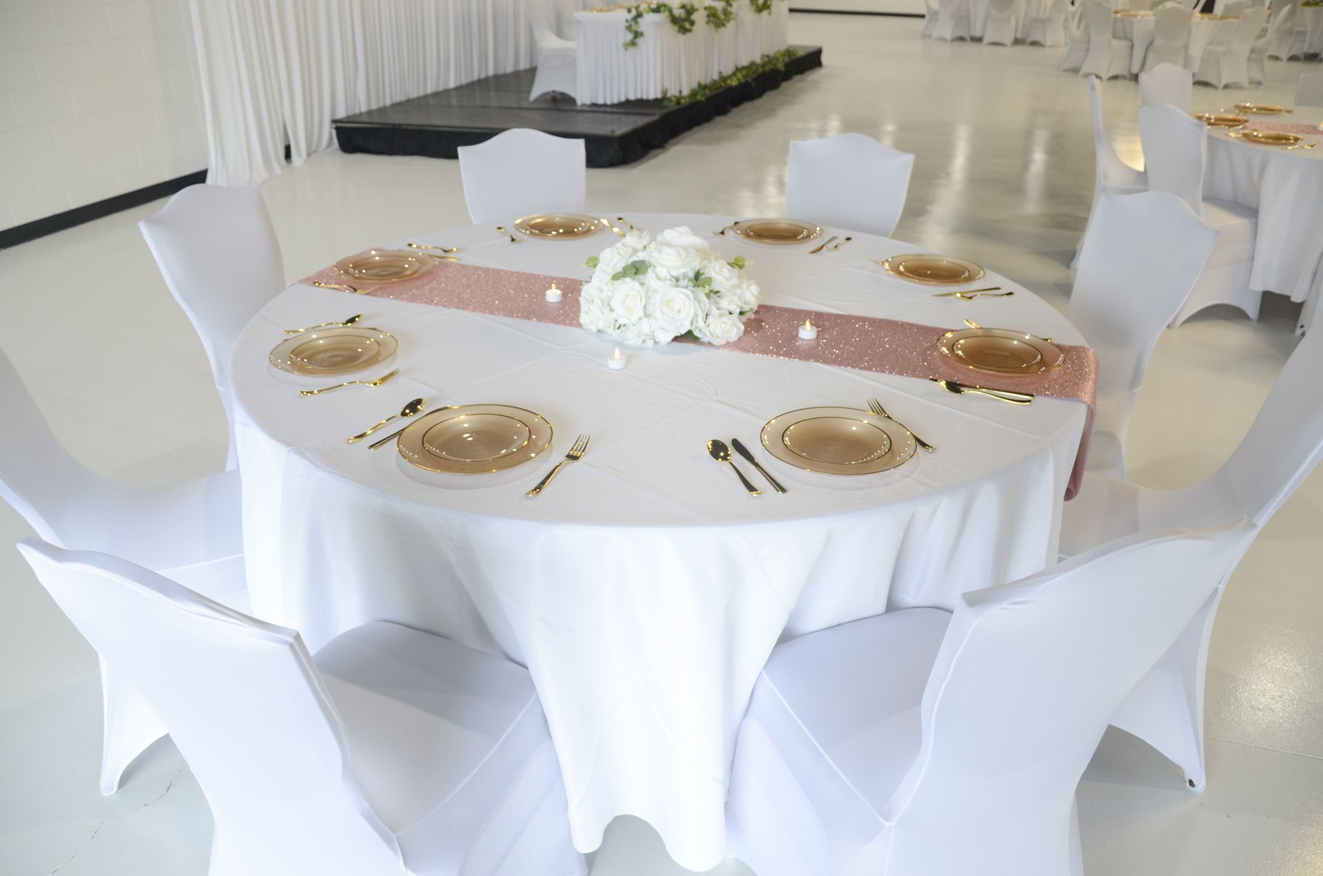 Round banquet table with white linen, gold place settings, and white chair covers in a bright event hall