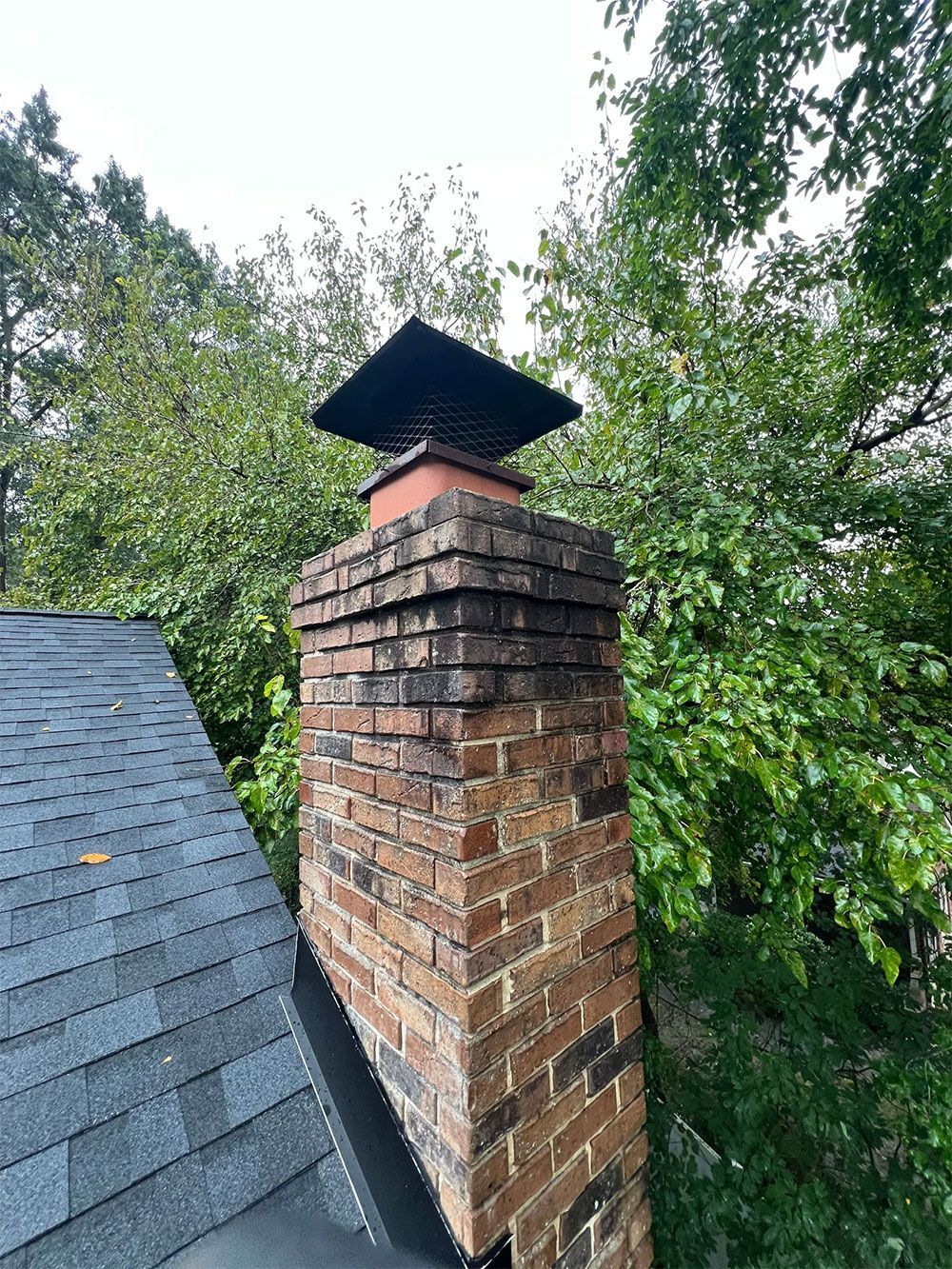 Brick chimney with a black cap on a dark roof, surrounded by green trees.