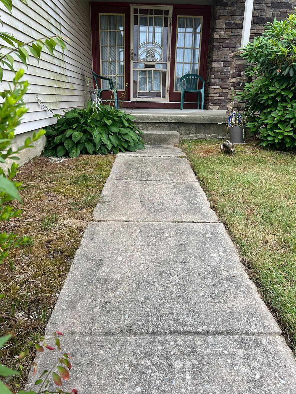 Concrete walkway leading to a front door with two chairs. Lush plants on either side.
