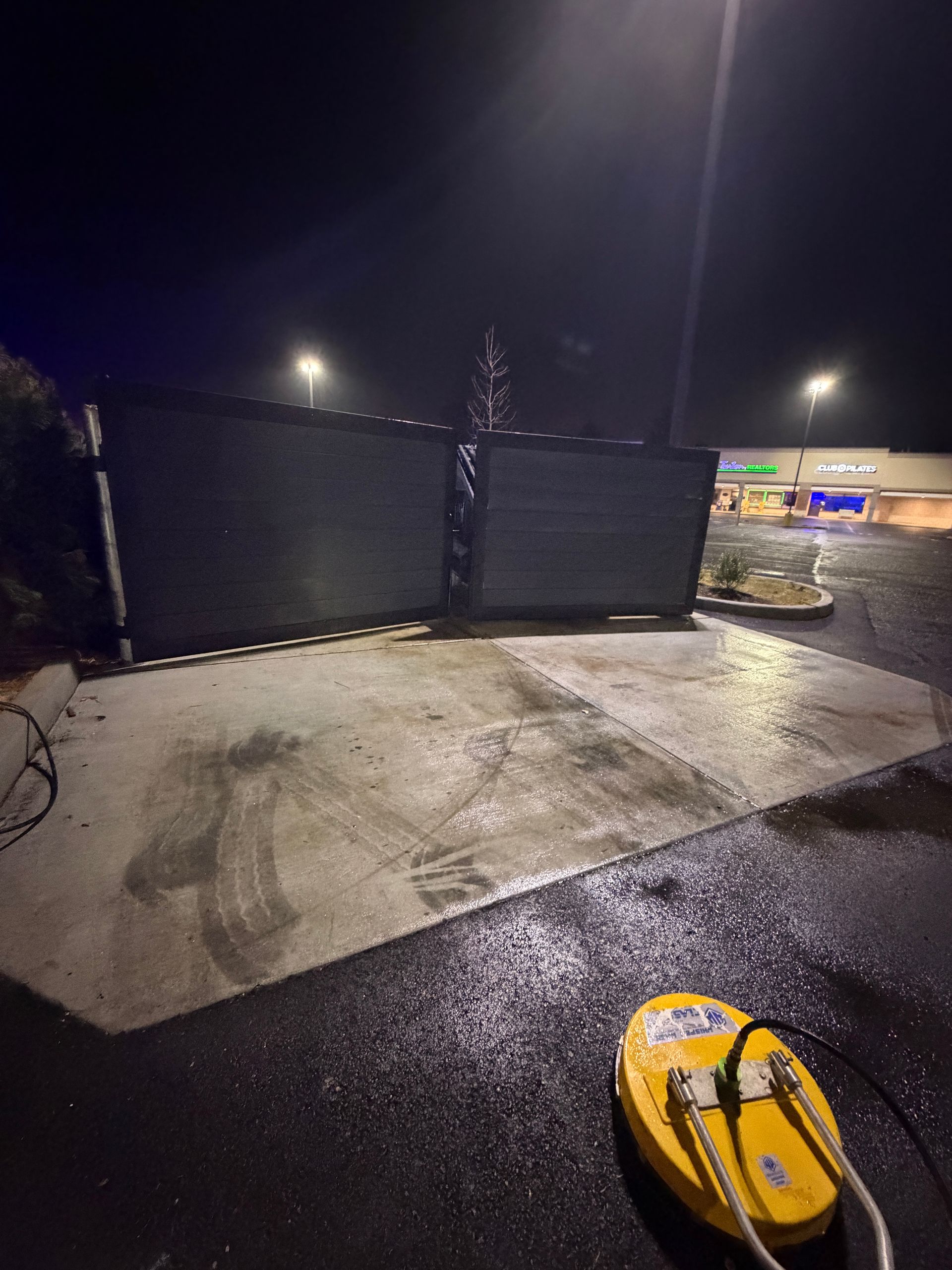Yellow pressure washer cleaning a concrete pad at night, near a dumpster and a store.