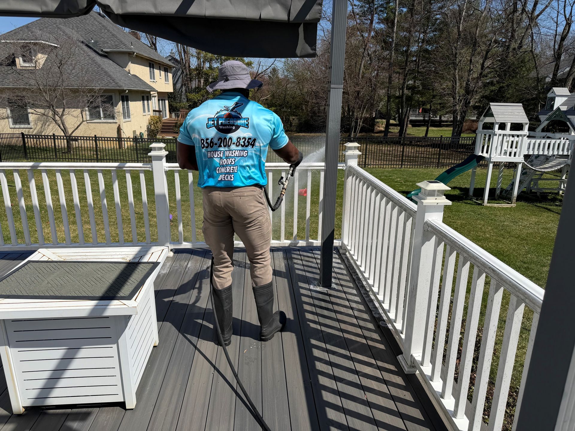 A person in a blue shirt and hat power-washes a deck, facing a green lawn with a playhouse in a residential backyard.