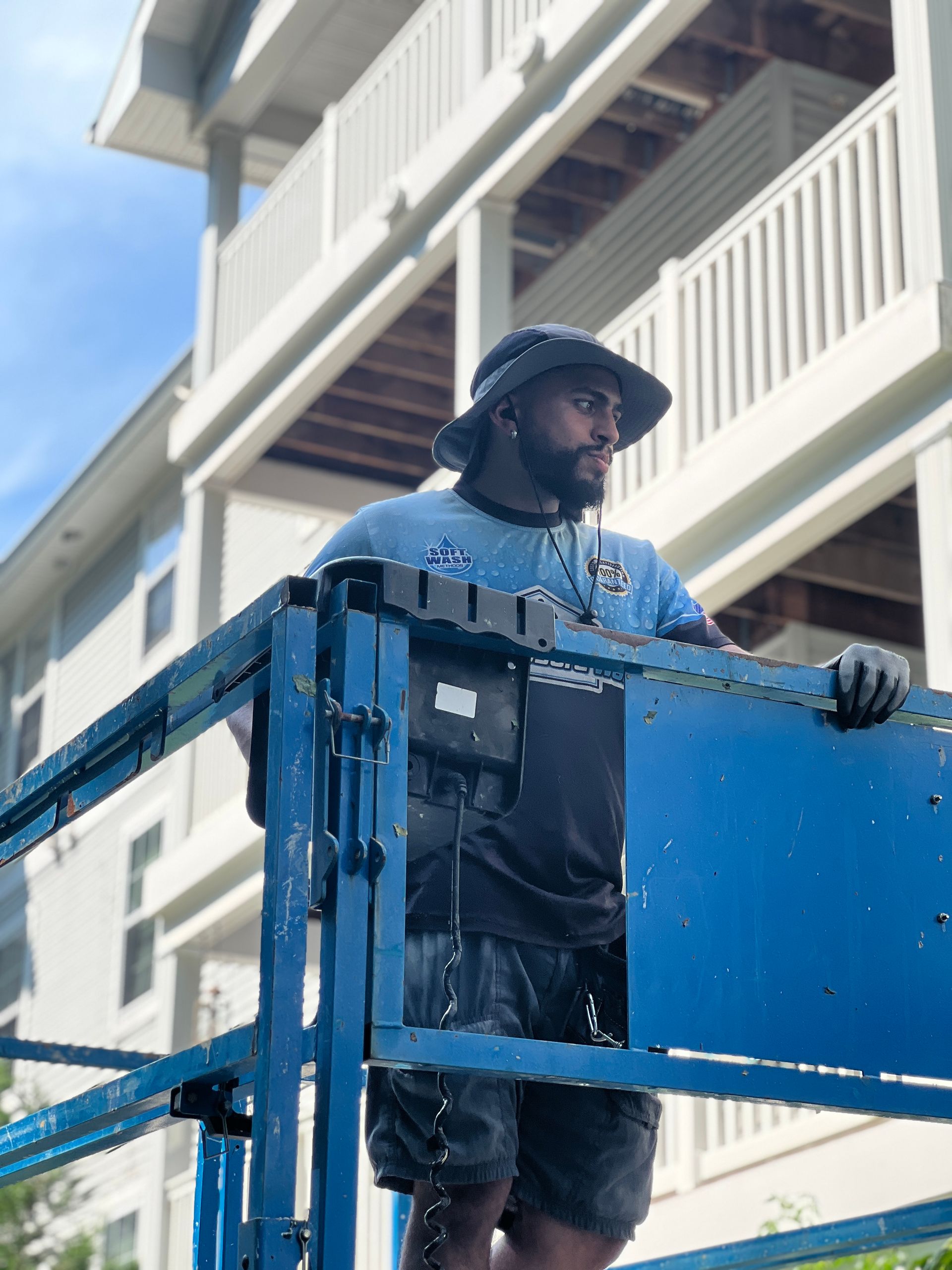 Person in lift operating equipment, looking up at building. Blue and grey tones, sunny outdoor setting.