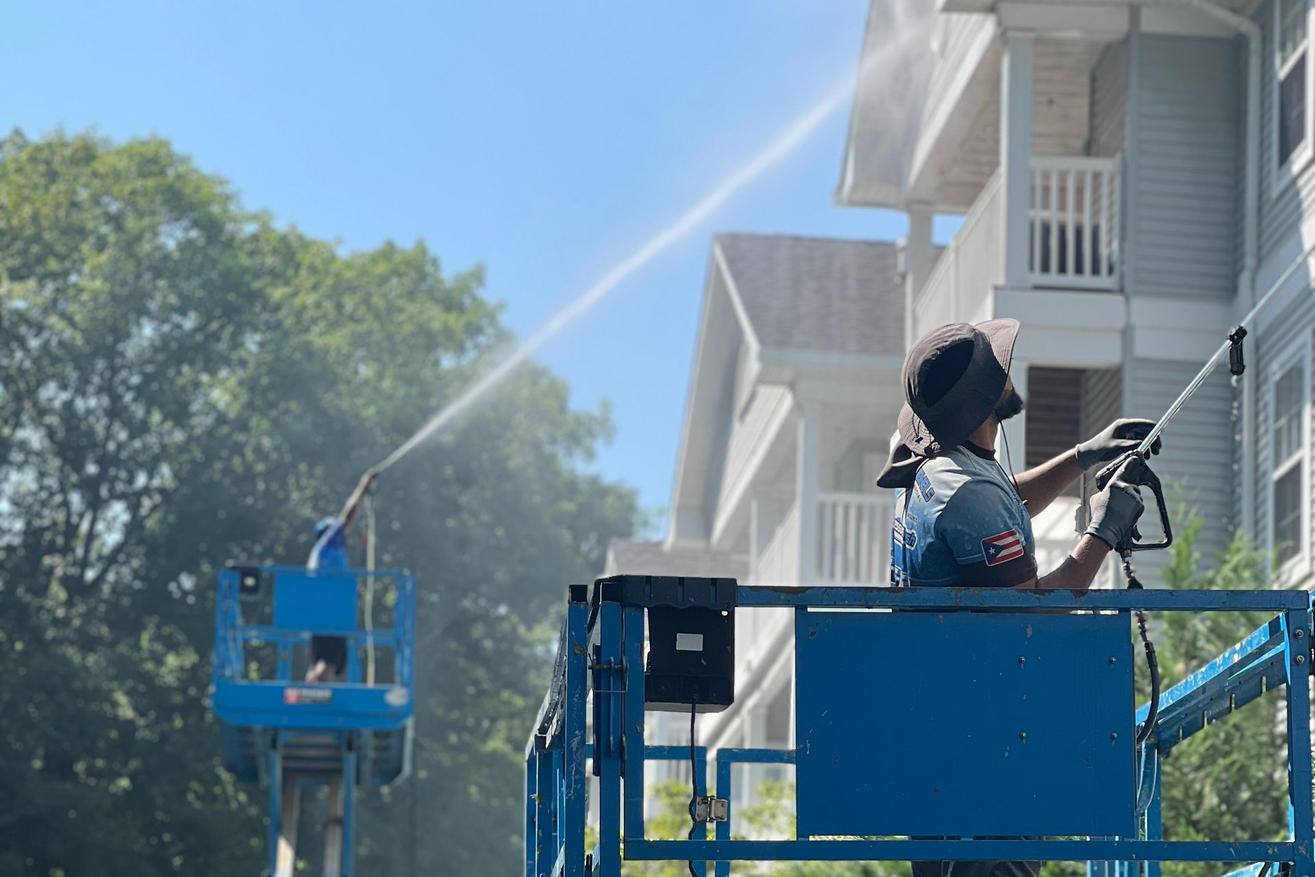 Two workers power washing a building from lifts; blue sky, sunny day.