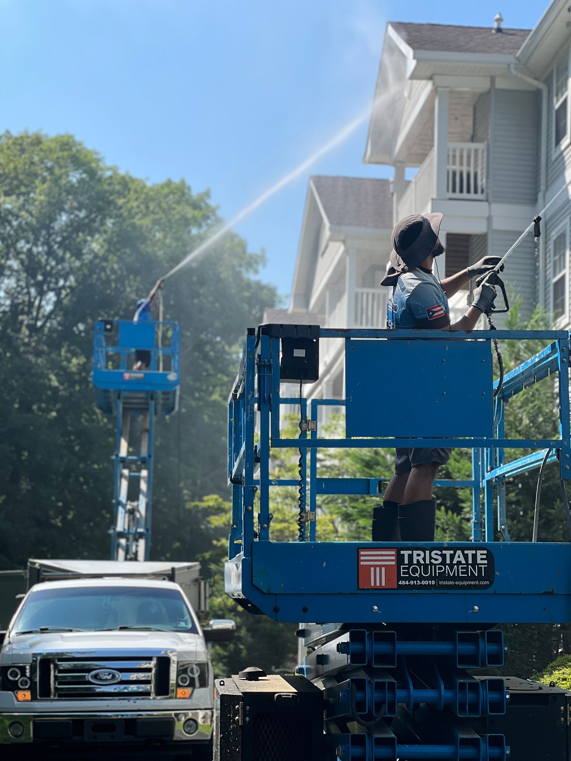 Two people using lifts to power wash a building exterior; one sprays from a lift. Truck parked in front.