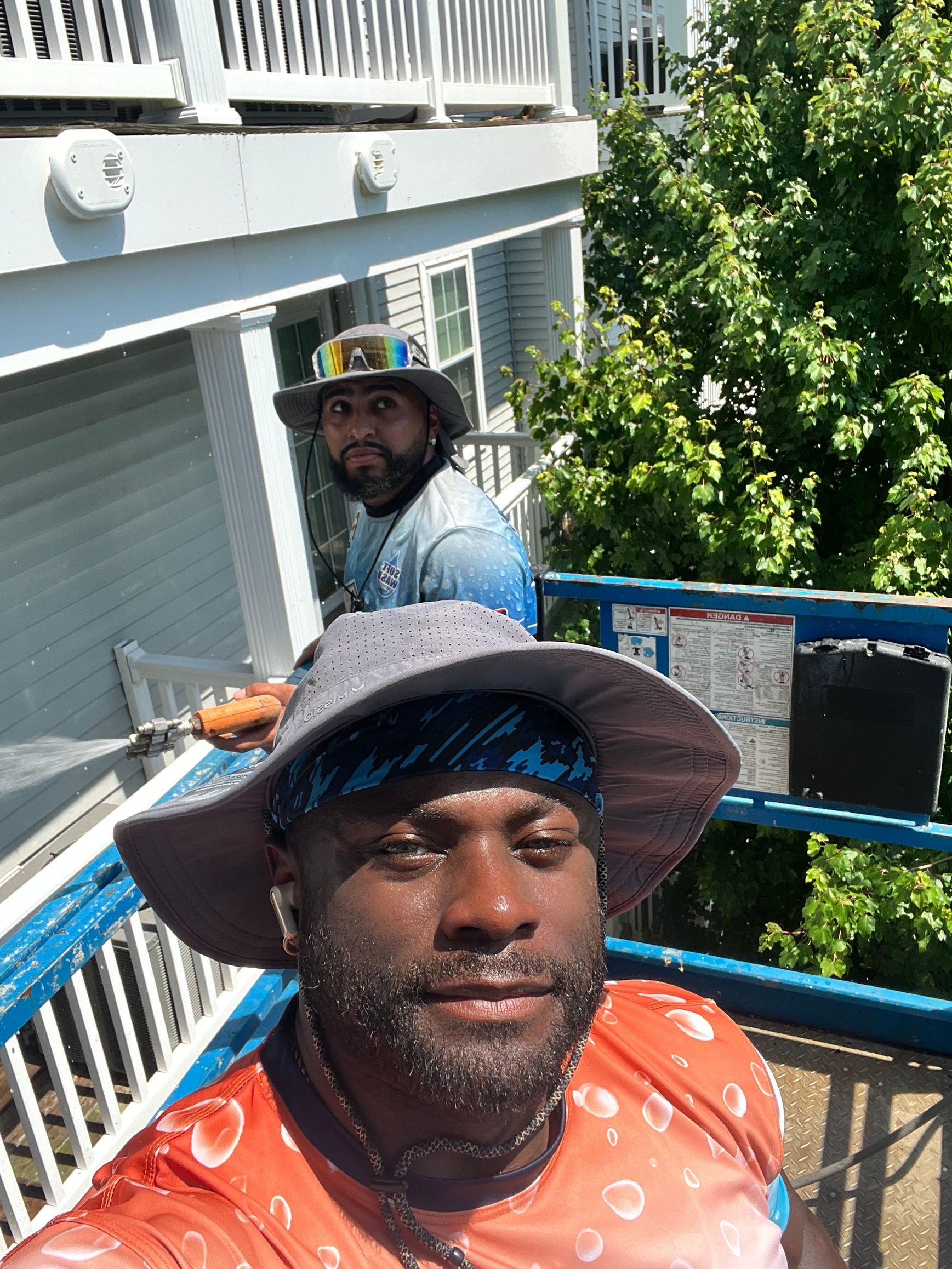 Two men on a lift; one takes selfie, the other points a hose. Building, foliage, and blue accents are visible.