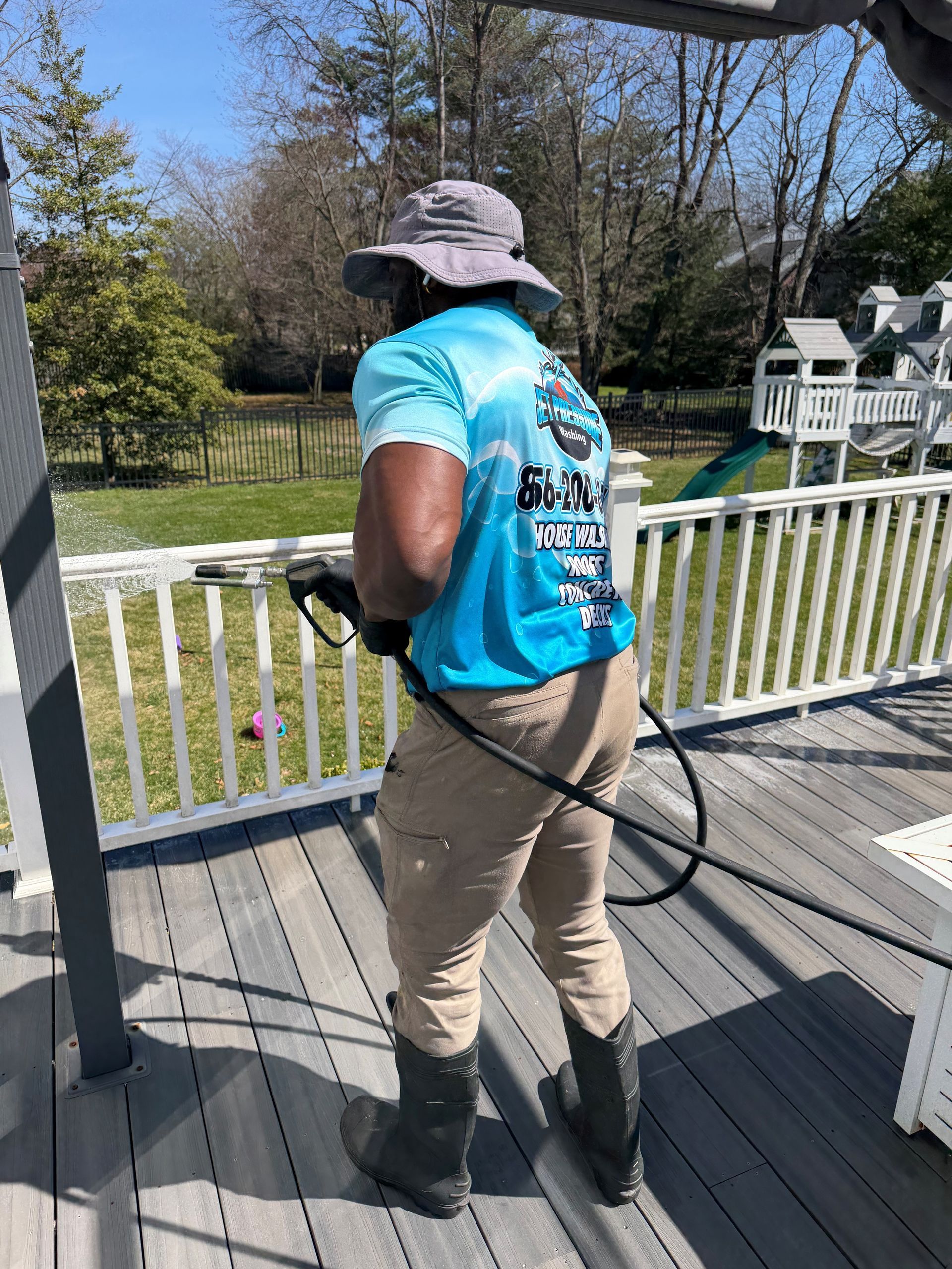 A person wearing a sun hat and work clothes uses a power washer to clean a gray deck on a sunny day.