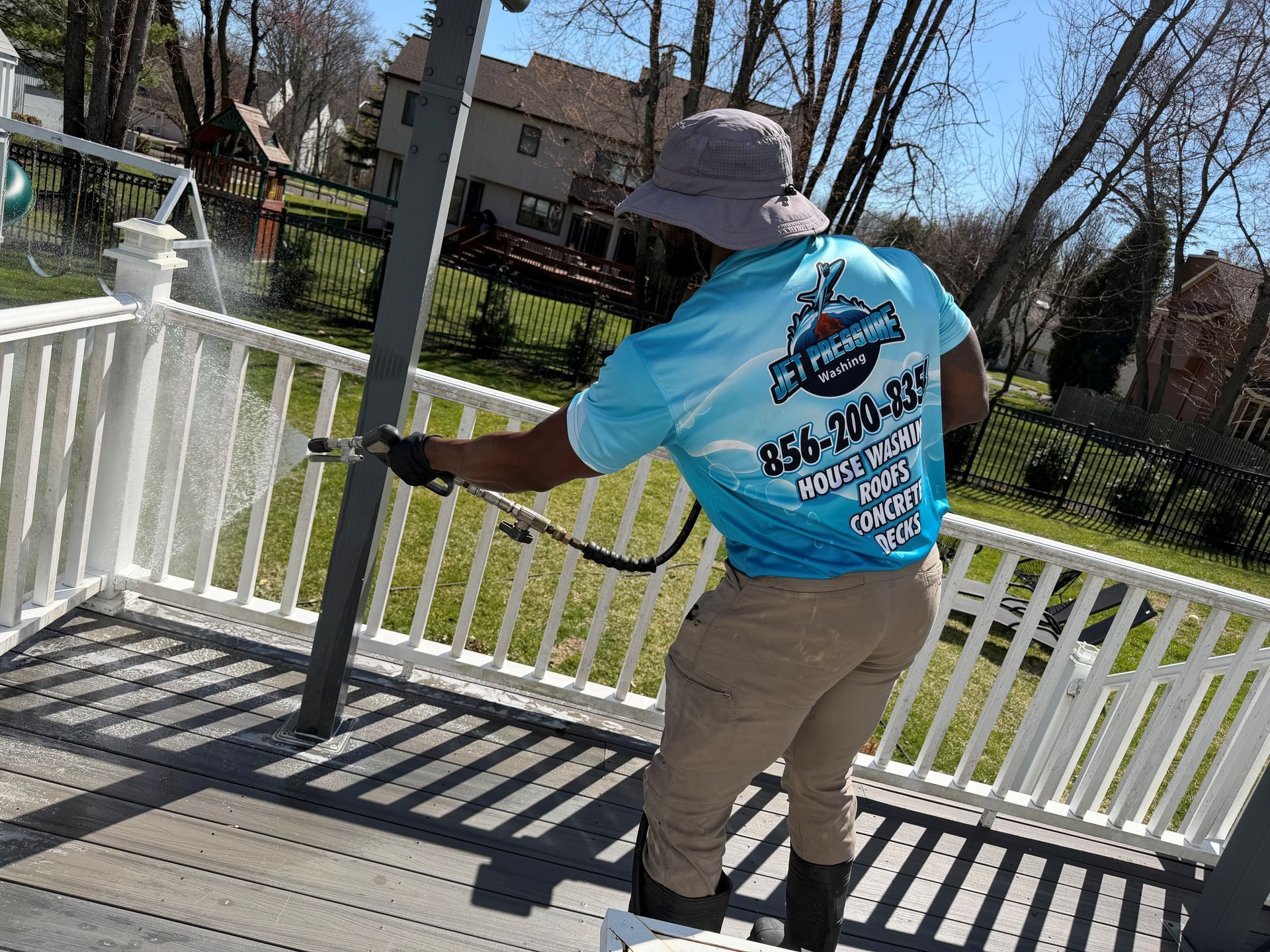 A worker in a blue branded shirt and bucket hat uses a pressure washer to clean a white deck railing outdoors.