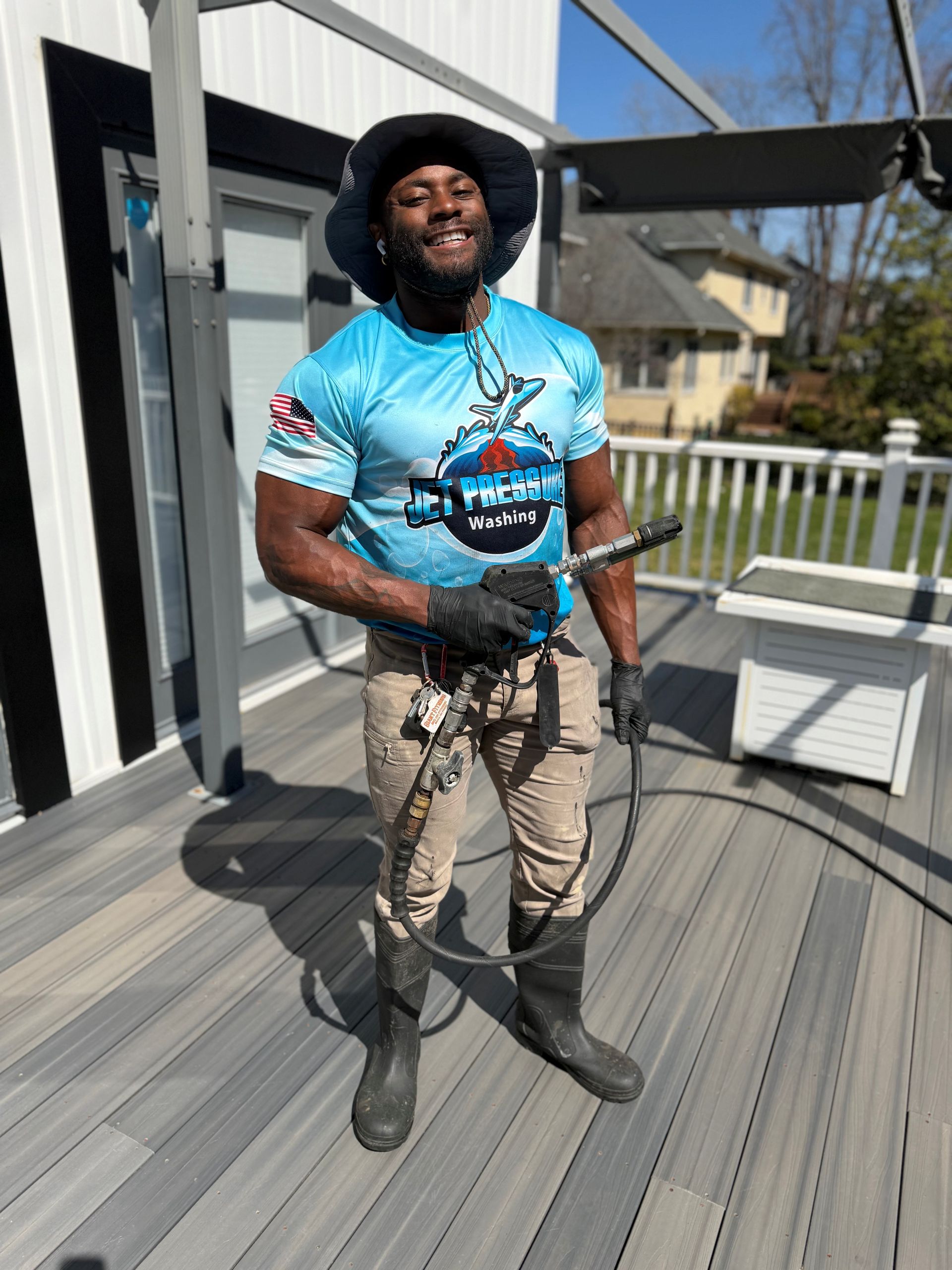 A smiling person in a blue graphic shirt, khaki pants, and boots holds a pressure washing wand on a grey composite deck.