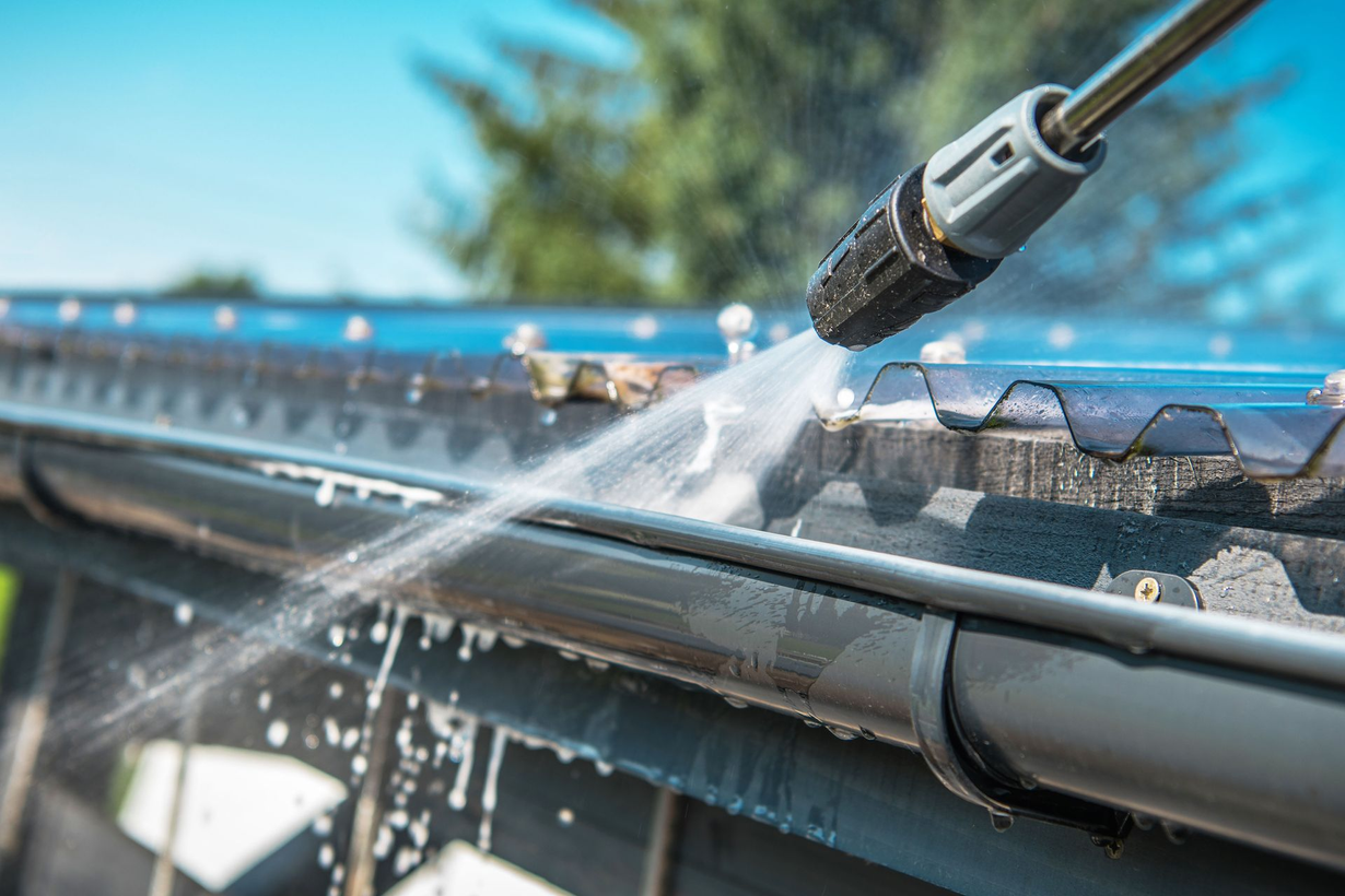 Power washer cleaning a dark gutter, clear sky, sunny day.