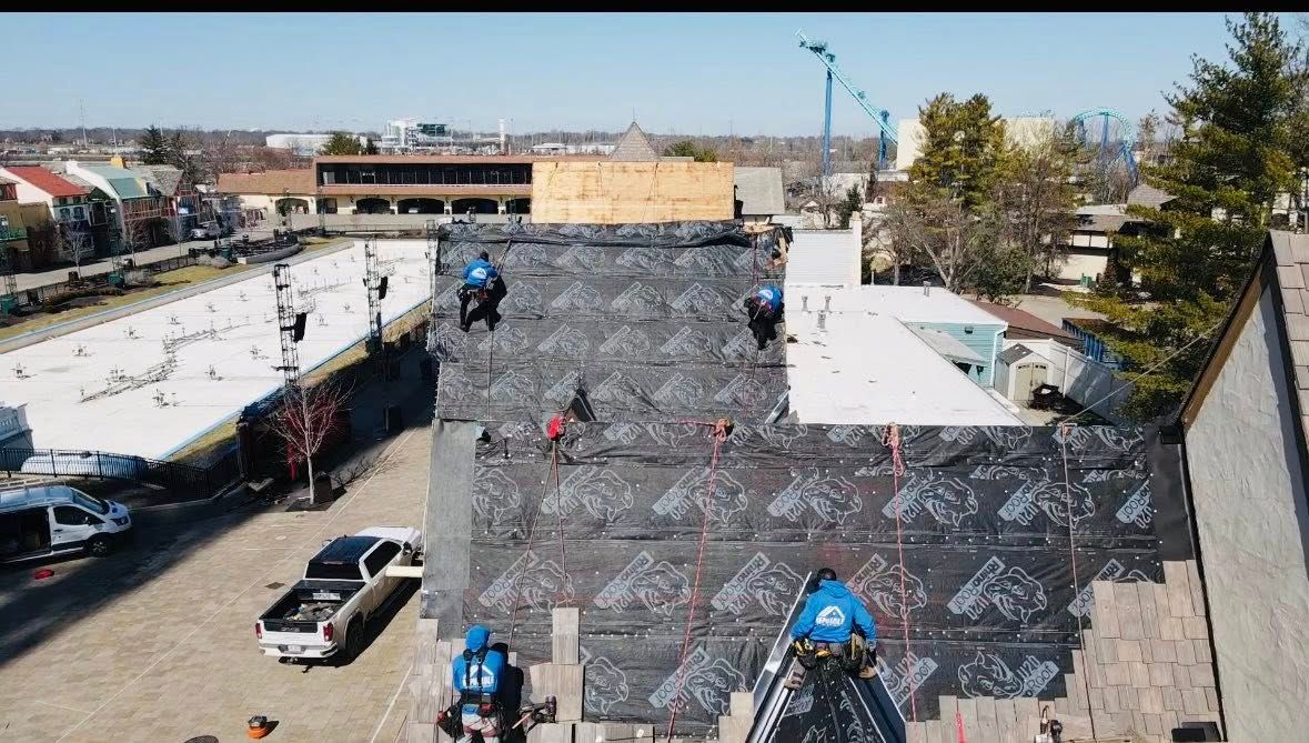 Roofers working on a building's roof with safety gear. Construction site with parked vehicles and urban backdrop.