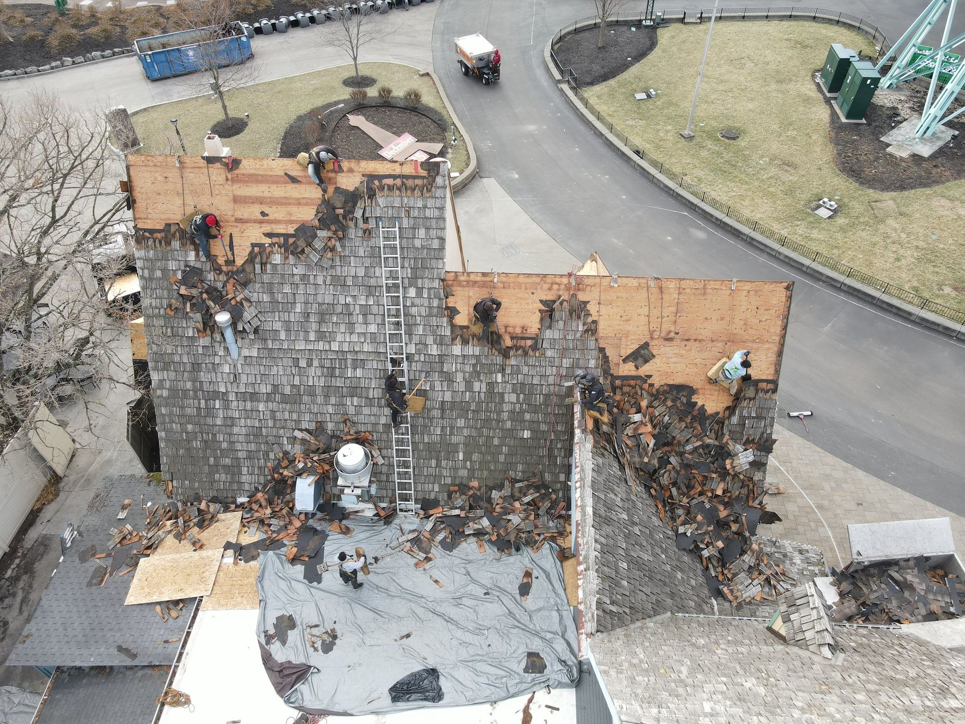 Roof of a building being repaired, workers on ladders, debris scattered, surrounded by asphalt and grass.