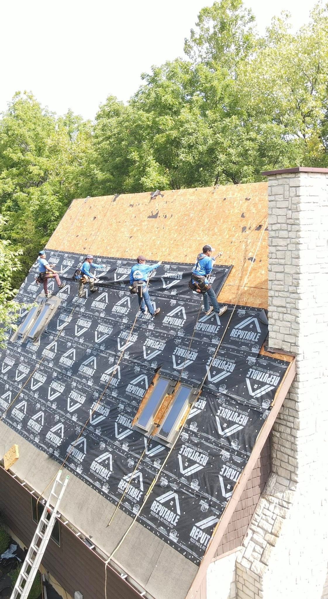 Roofers installing roofing material on a house, trees in the background, a ladder on the side.
