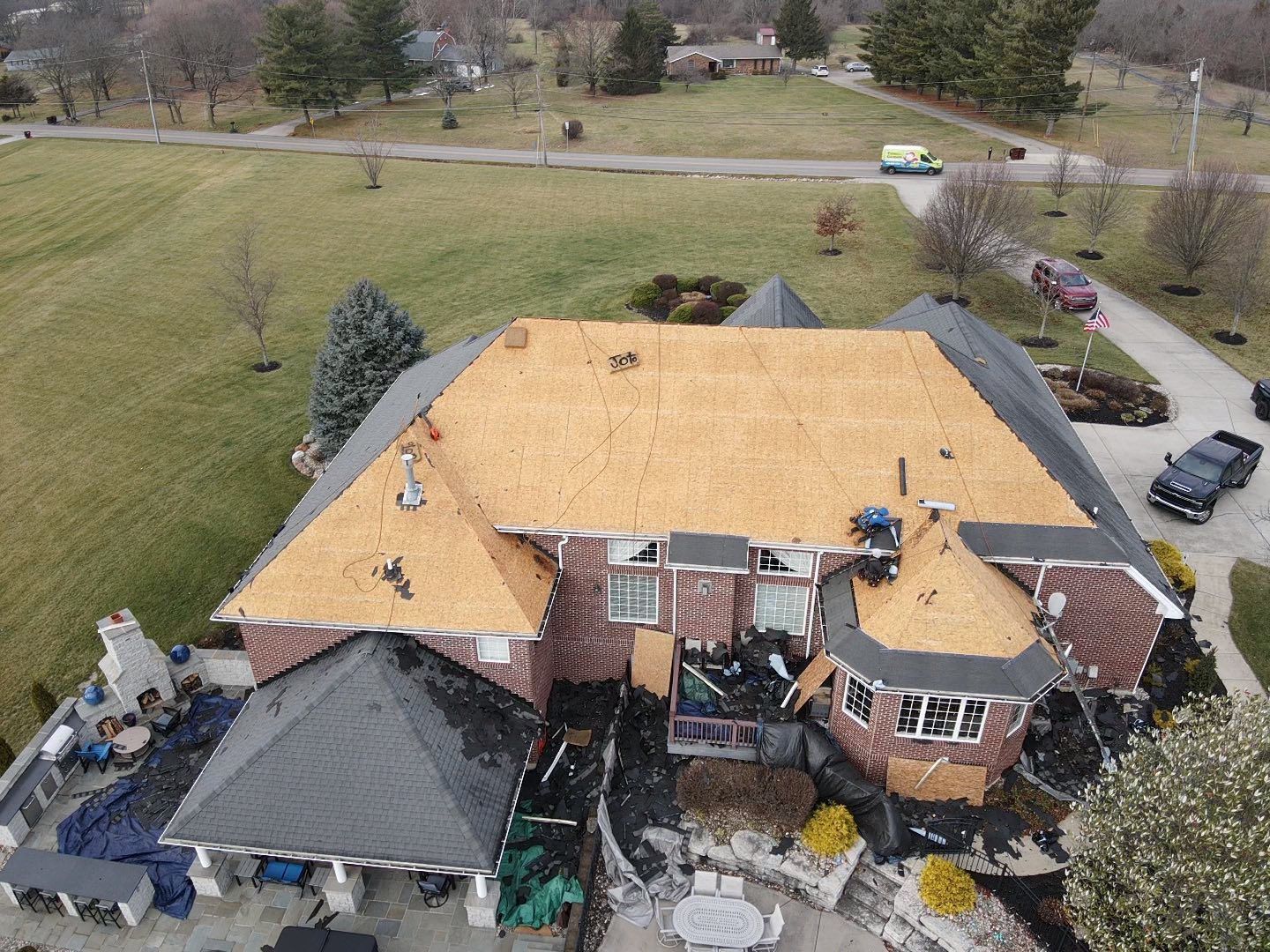 Aerial view of a house with roofing in progress. Parts of the roof are covered and uncovered, with materials on the ground.