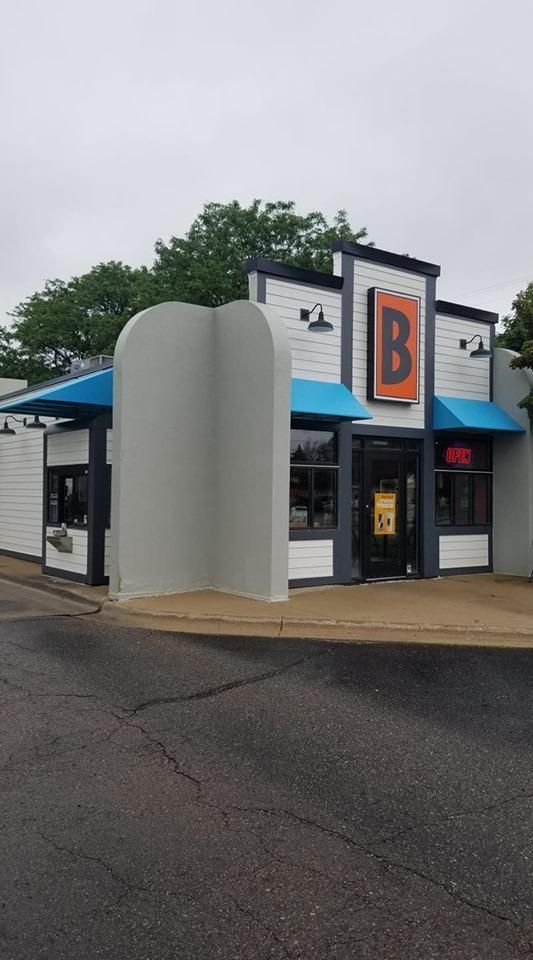 Small white-and-black storefront with a large orange “B” sign, seen on a wet street under overcast skies.