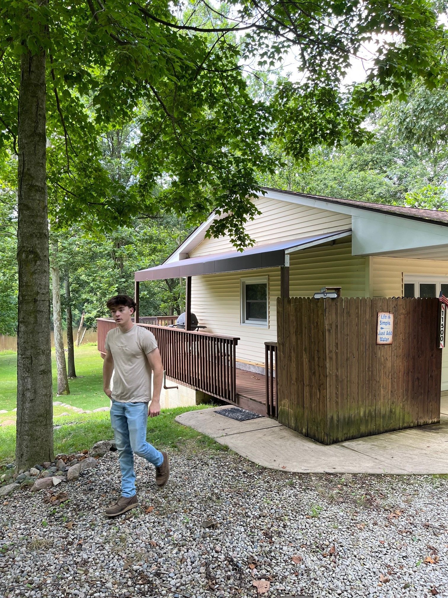 Person walking near a small building in a wooded area, with gravel ground and trees overhead