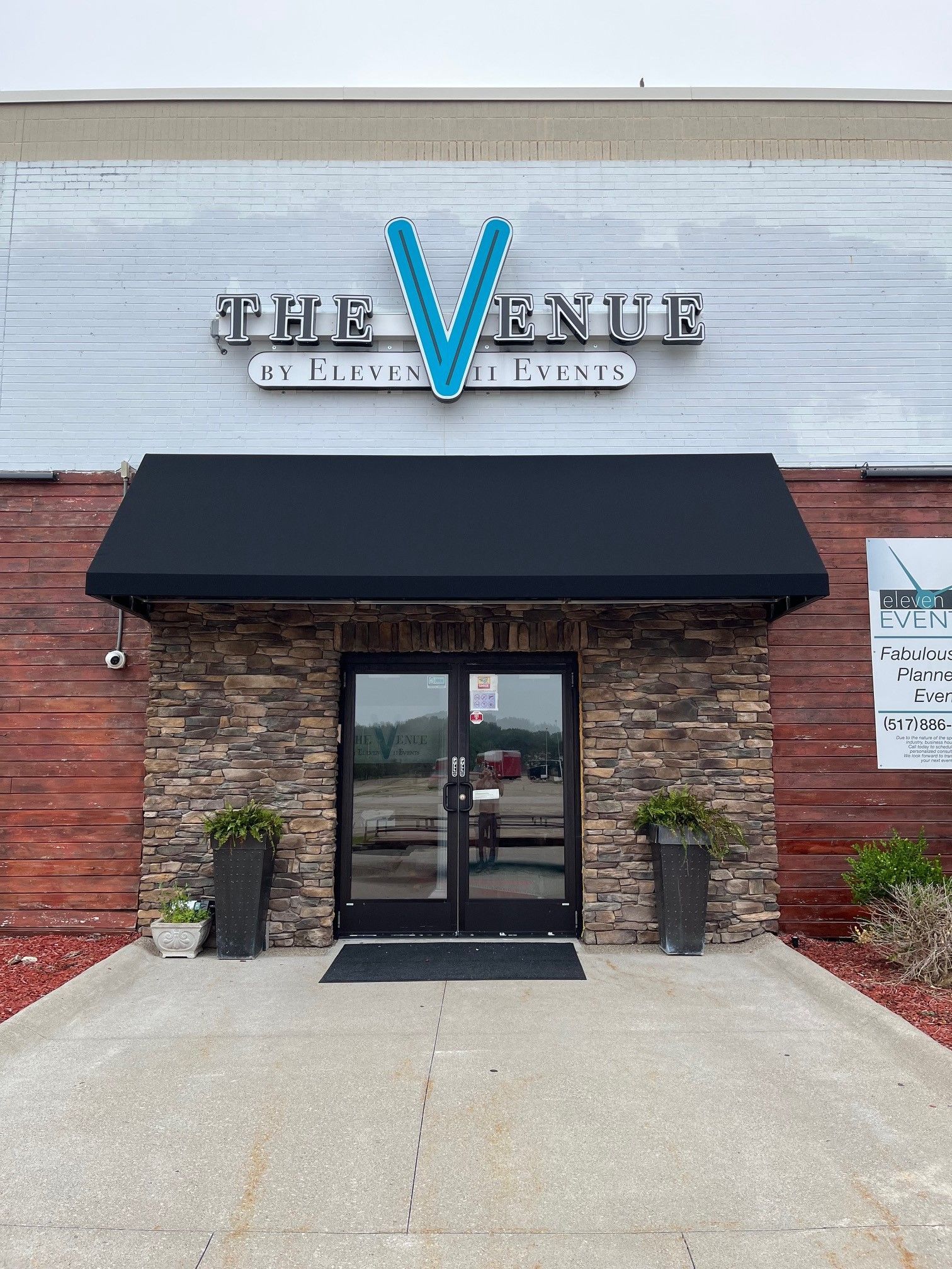 Storefront entrance with black awning, stone facade, and blue “V” sign above glass doors