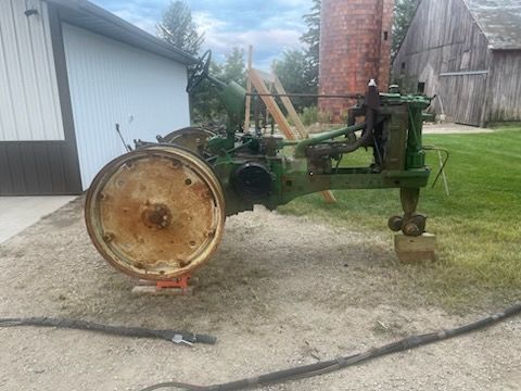 Green John Deere tractor, missing parts, outdoors. Rusty wheel, red brick silo, and barn in background.
