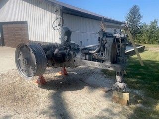 An old gray tractor sits on jacks outdoors near a white building on a sunny day.