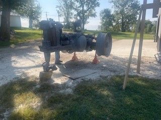 An old machine raised on jacks outside on a grassy area near trees and a building.