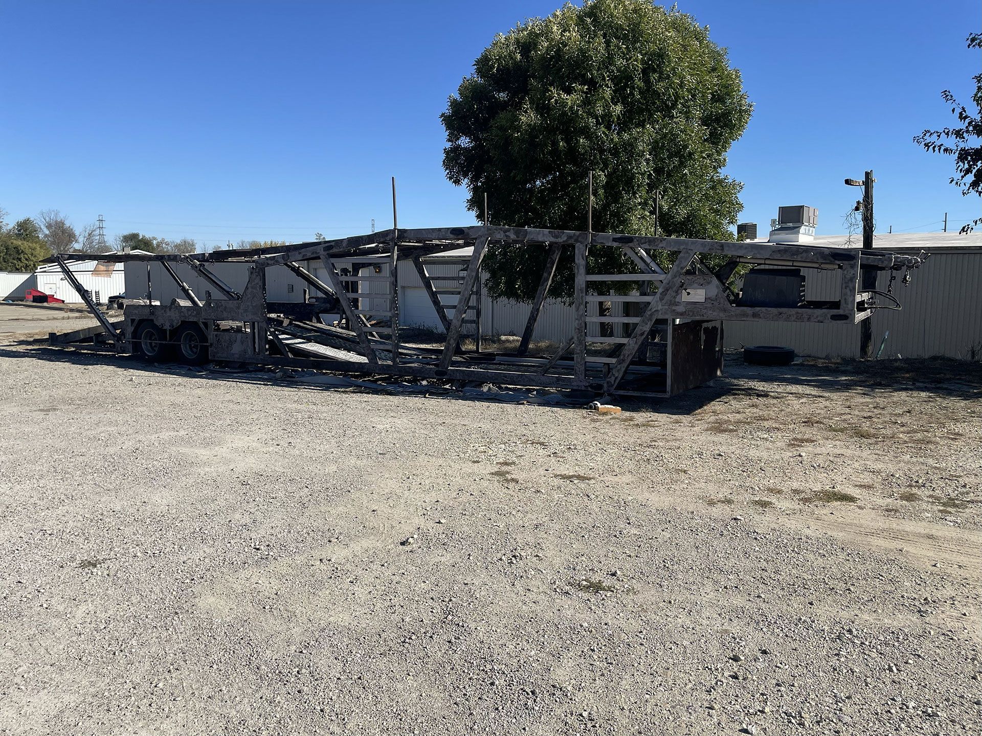 Empty car hauler trailer parked on gravel in front of a fence and building.
