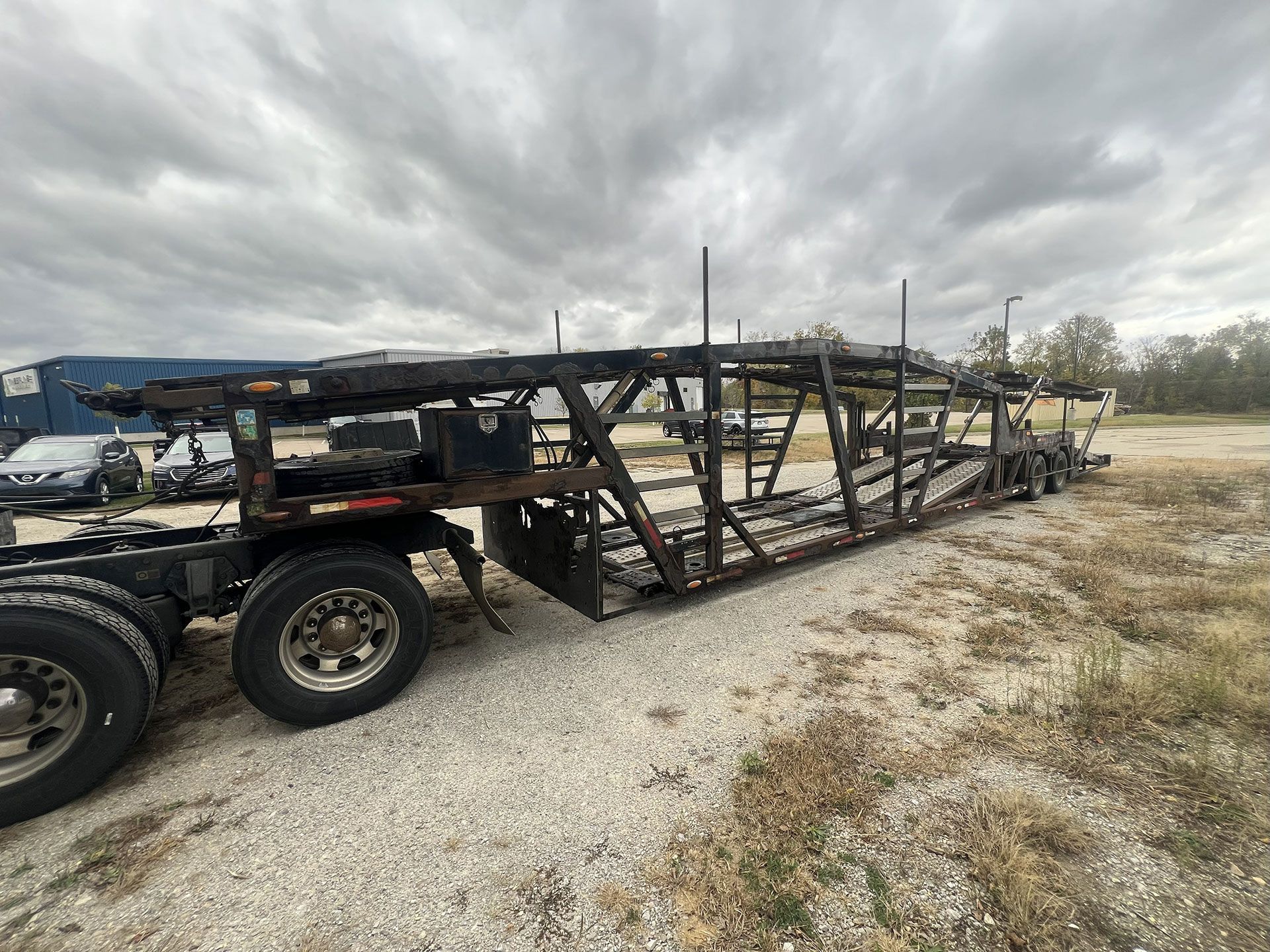 A burnt, empty car hauler trailer sits on gravel under a cloudy sky.