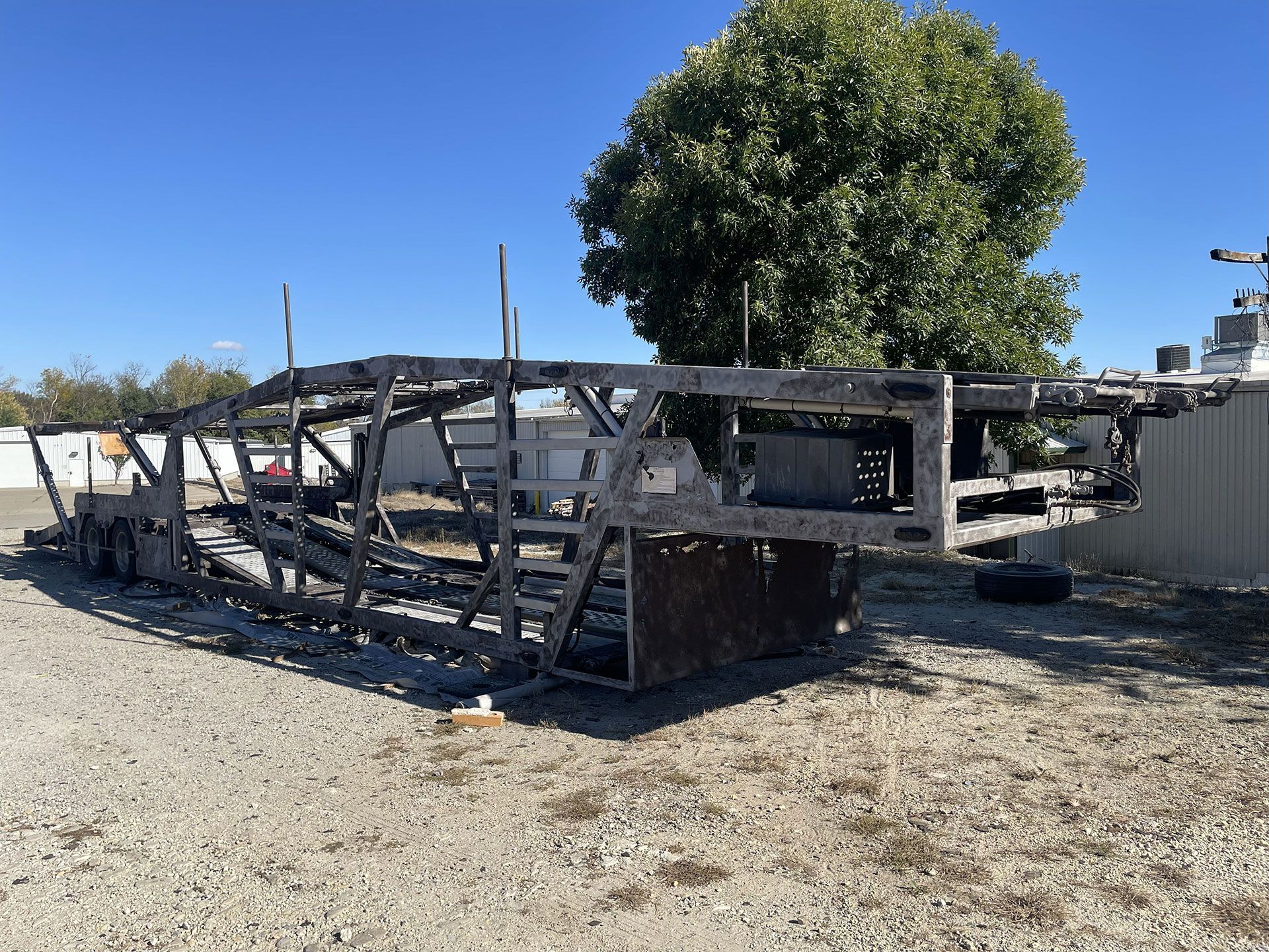 Burnt wooden trailer frame on gravel, with a tree and blue sky in the background.