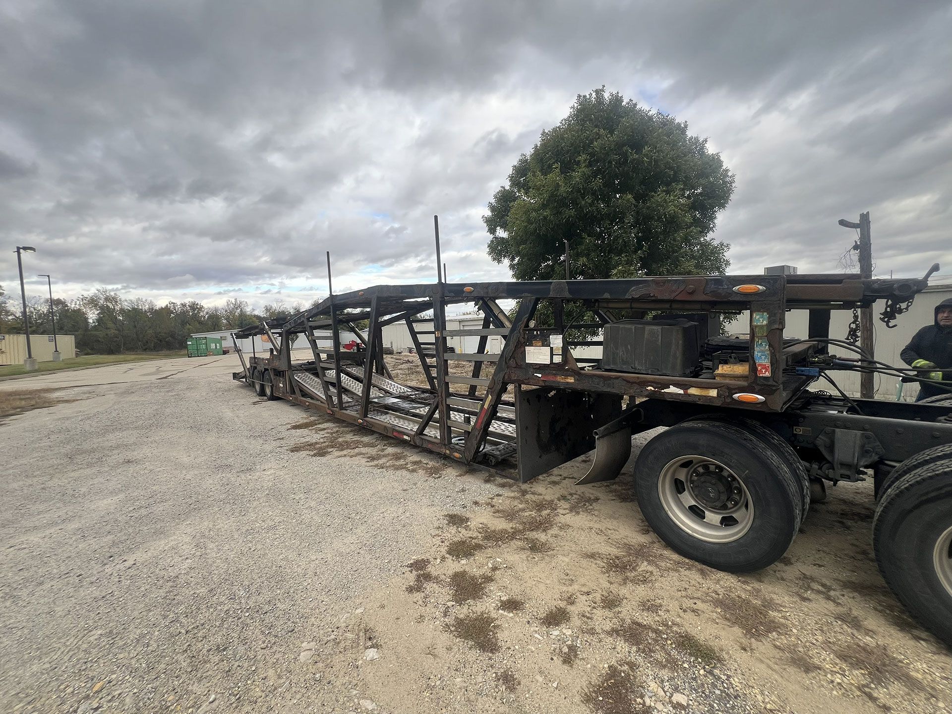 Burnt-out car hauler trailer on gravel lot under cloudy sky.