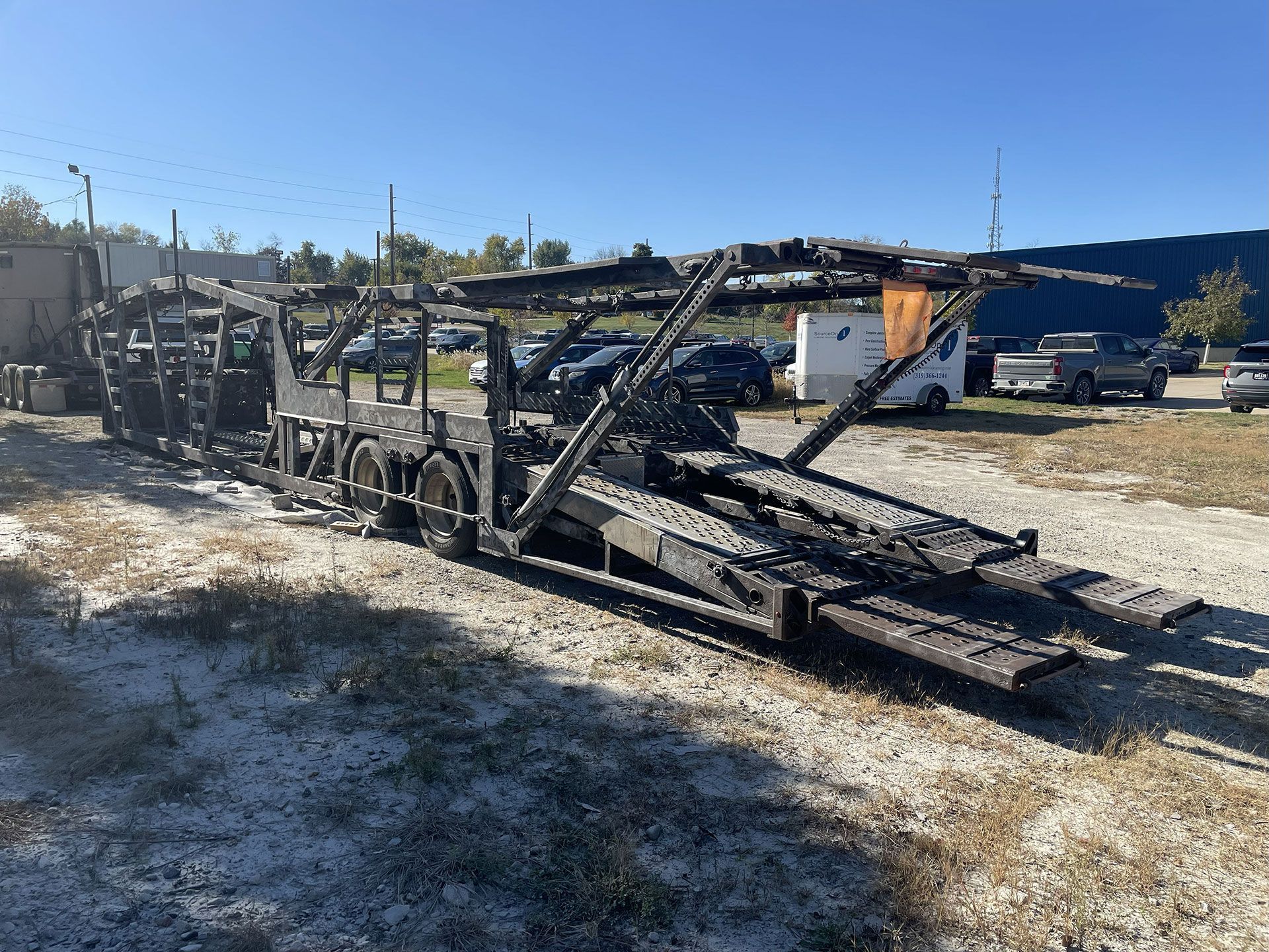 Burned-out car carrier trailer on a grassy lot, charred remains against a blue sky.