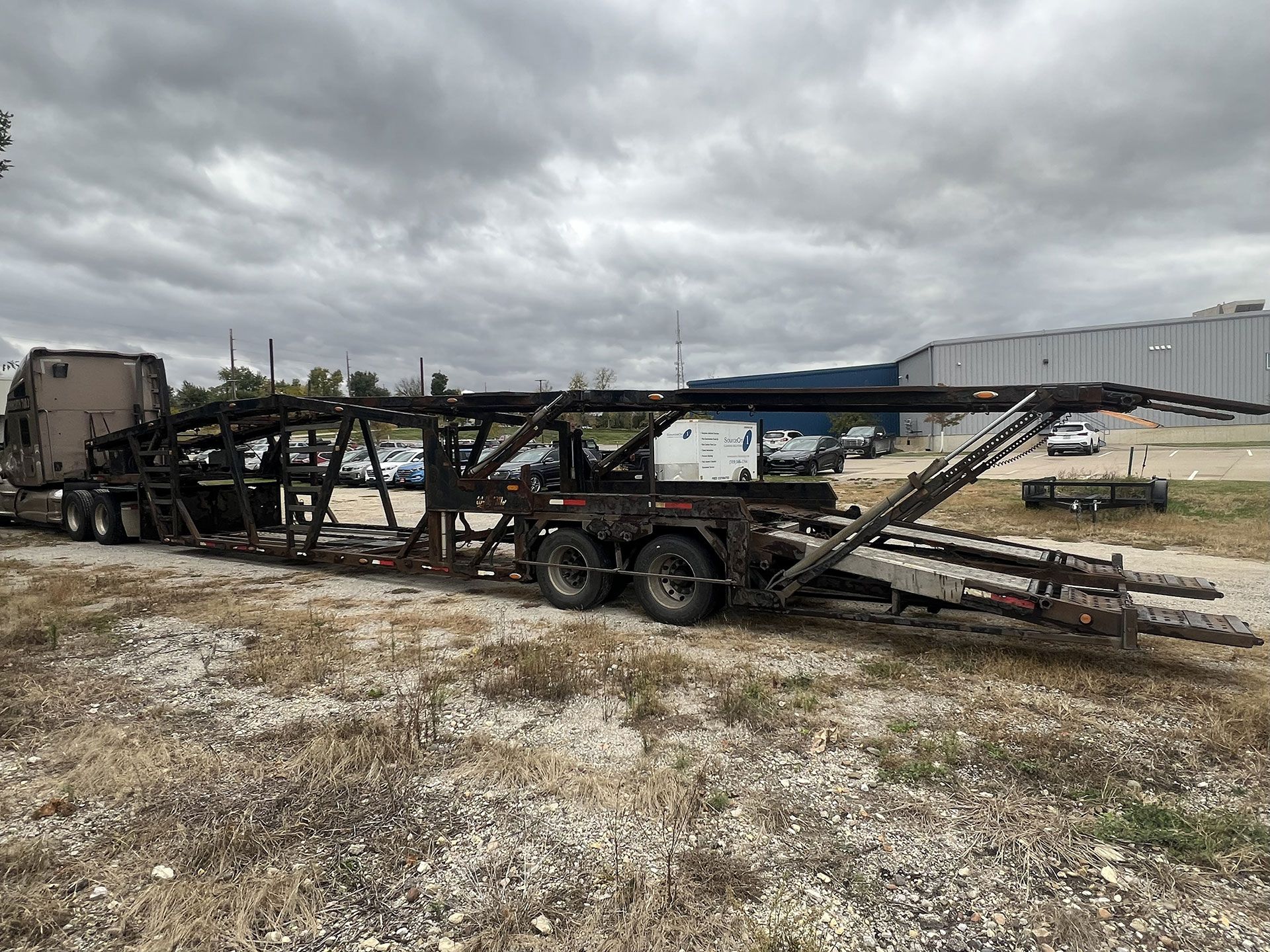 A car hauler truck parked on a grassy area, overcast sky. The trailer is empty.