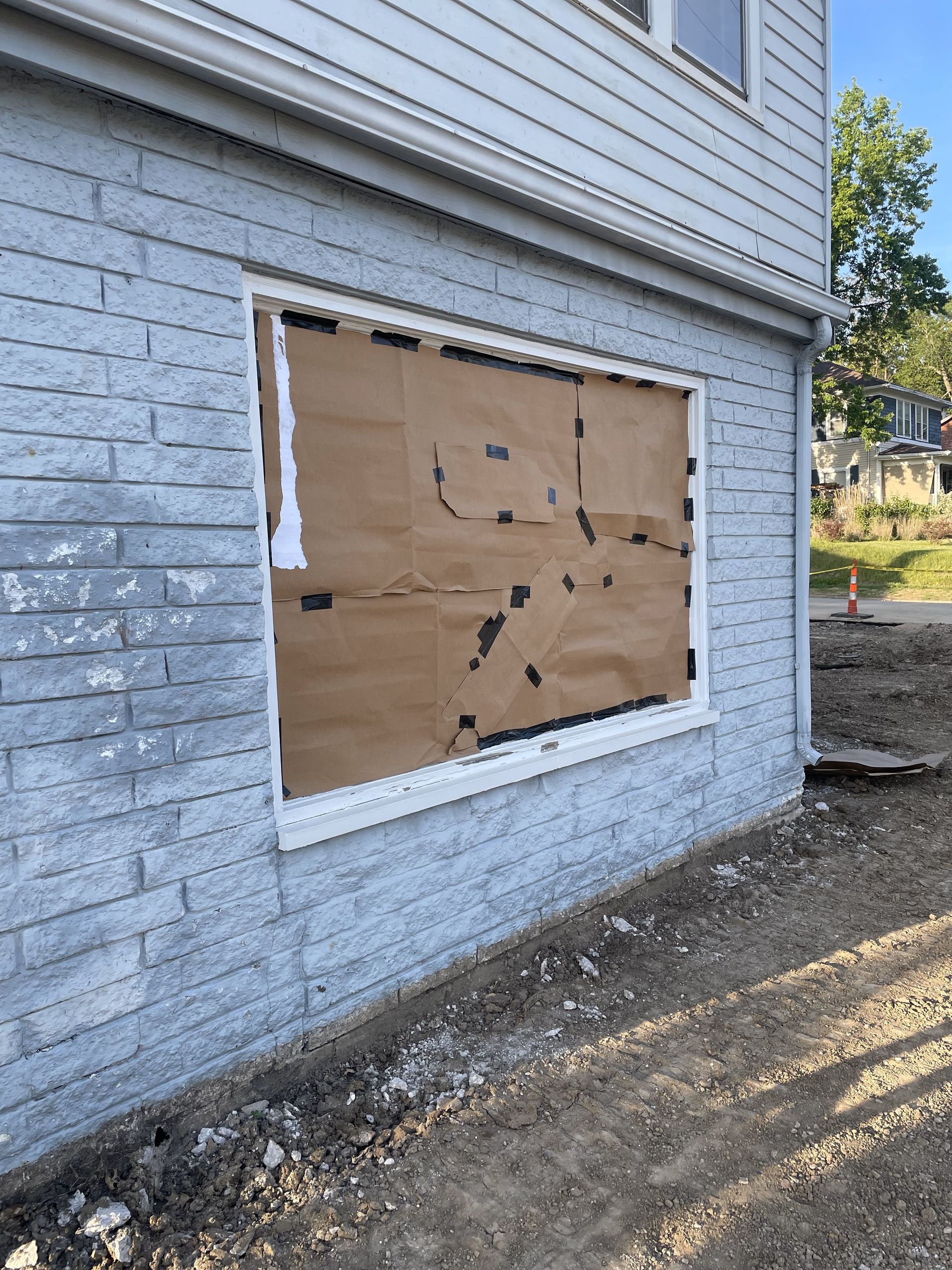 Exterior view of a building with a boarded-up window covered in brown cardboard and black tape, set against a backdrop of construction and blue siding.