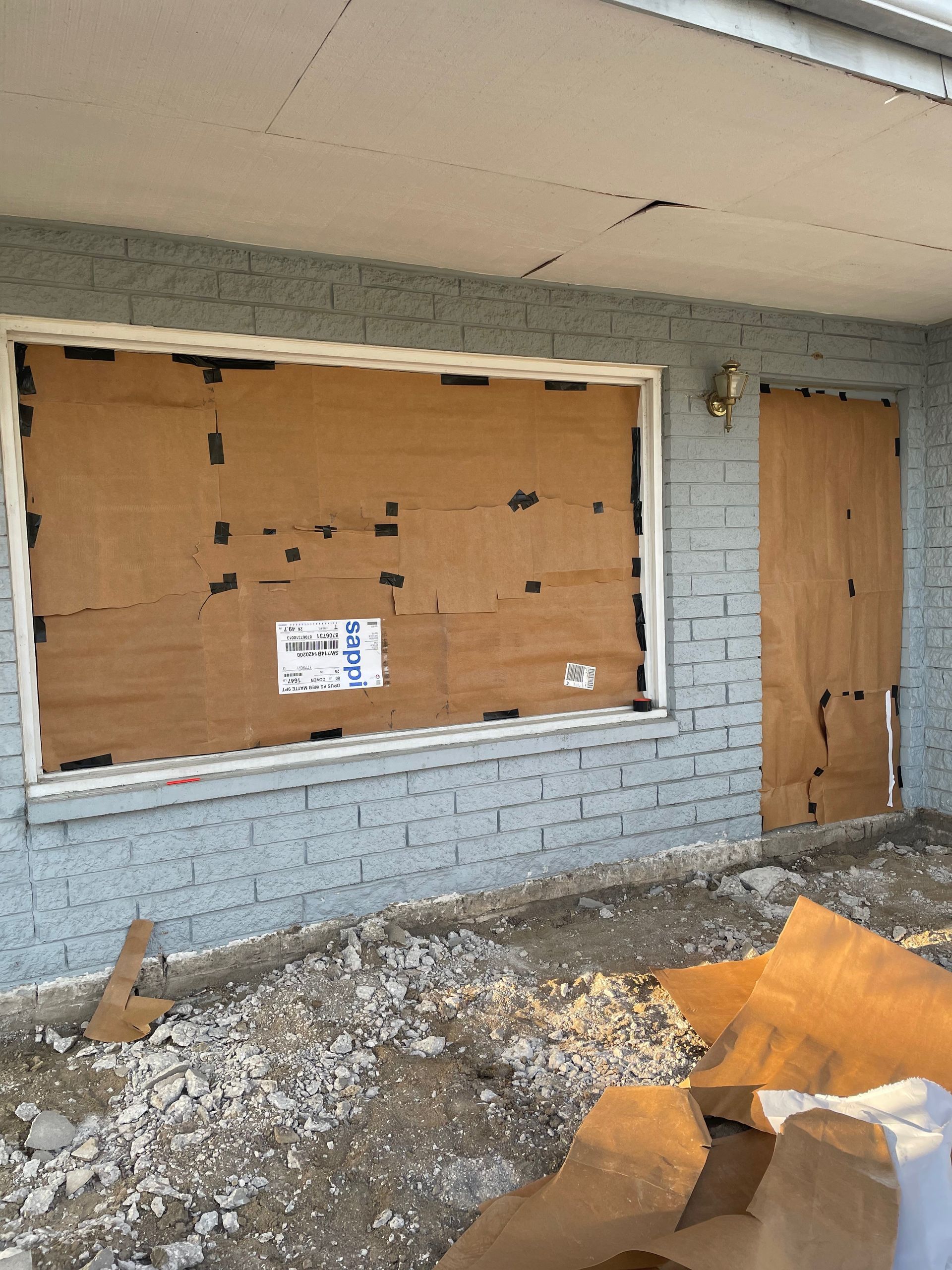 A building with a boarded-up window and door. The building has gray brick and a dirt yard.