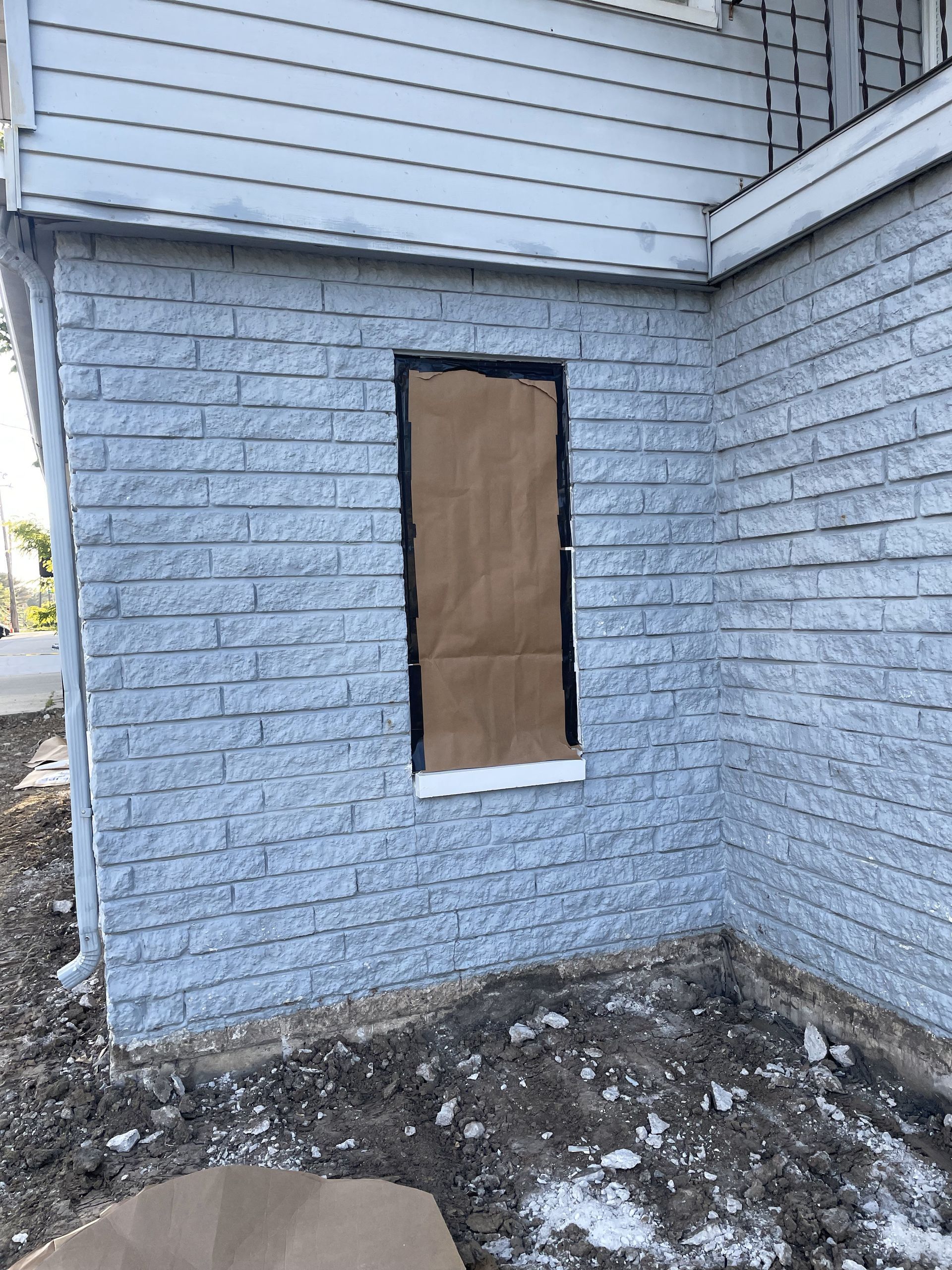 Exterior wall of a house with blue brick-like siding, a boarded-up rectangular opening, and exposed dirt at the base.