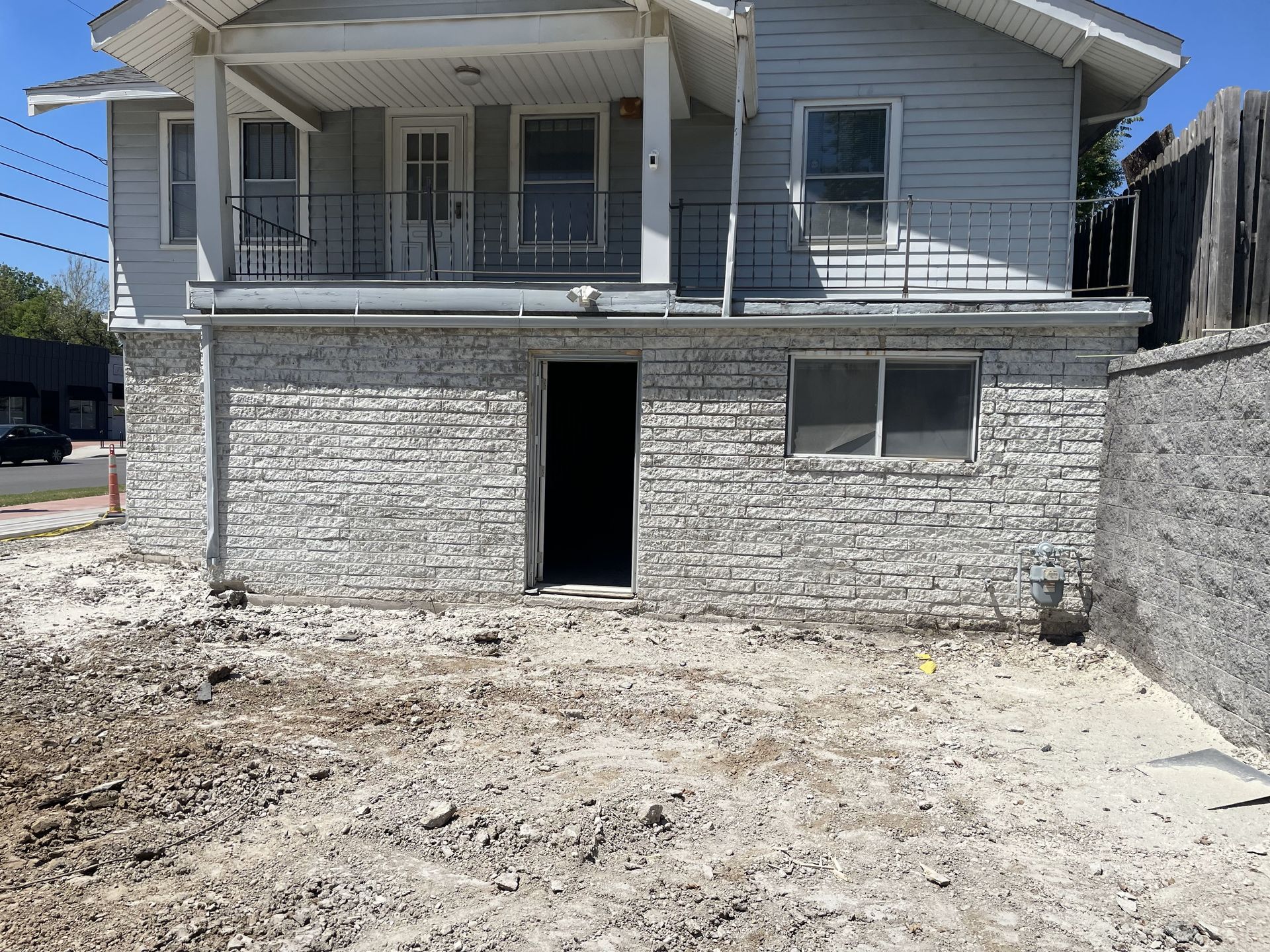 A two-story house with a stone-clad lower level and a porch. The yard is unpaved with exposed dirt and debris.