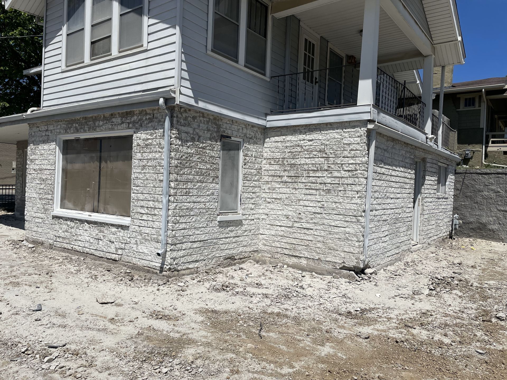 Corner of a house with light gray stone facade on lower level, gray siding above. Windows and gutters visible, gravel ground.