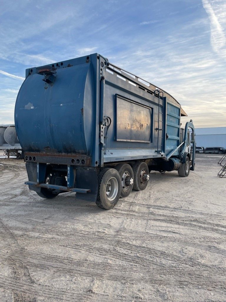 A blue garbage truck is parked in a dirt lot