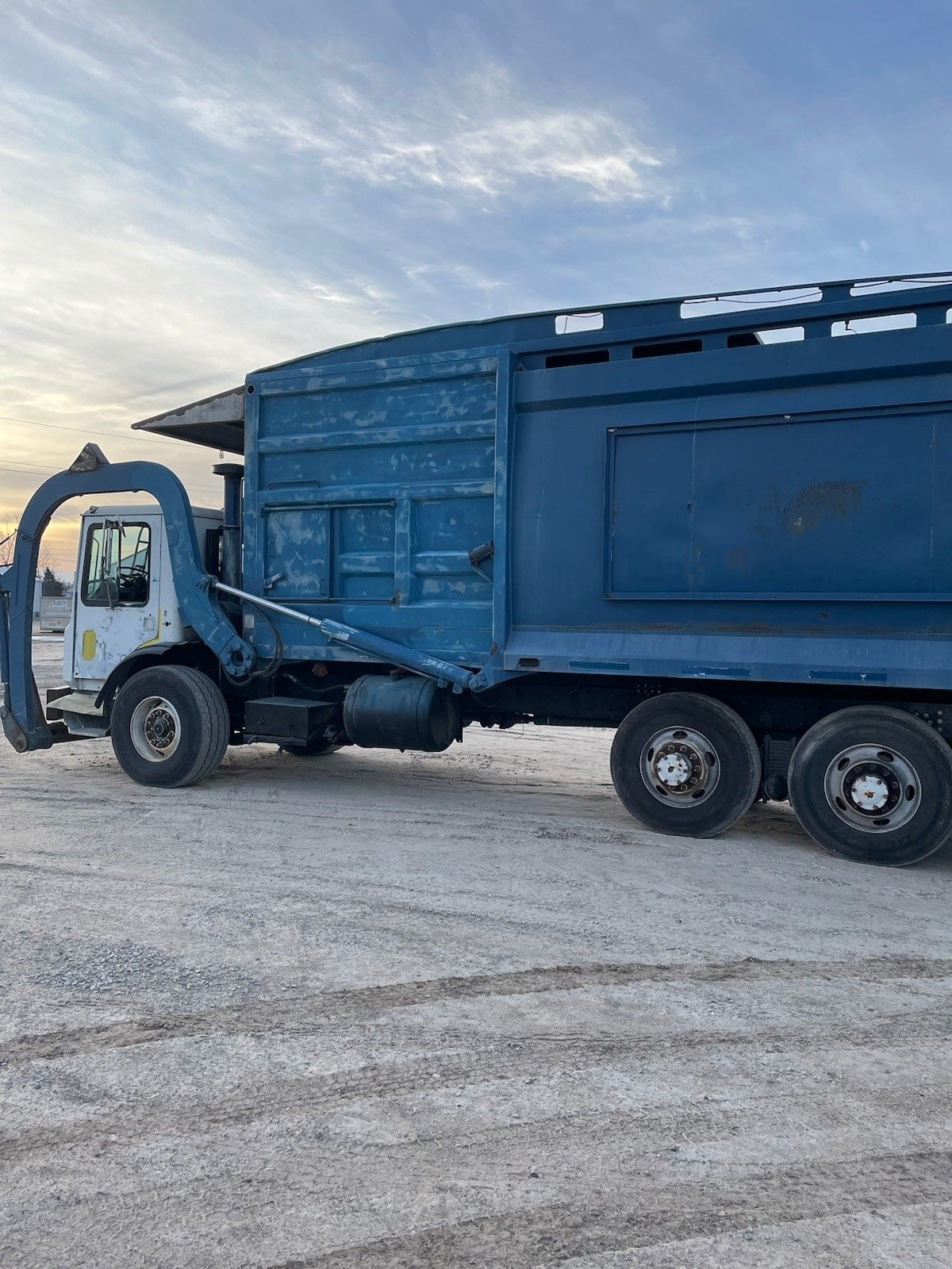a blue garbage truck is parked in a dirt field