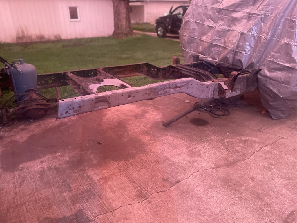 A vehicle frame sits on a red-tinted concrete surface in a yard. A tarp partially covers a large structure behind the frame.