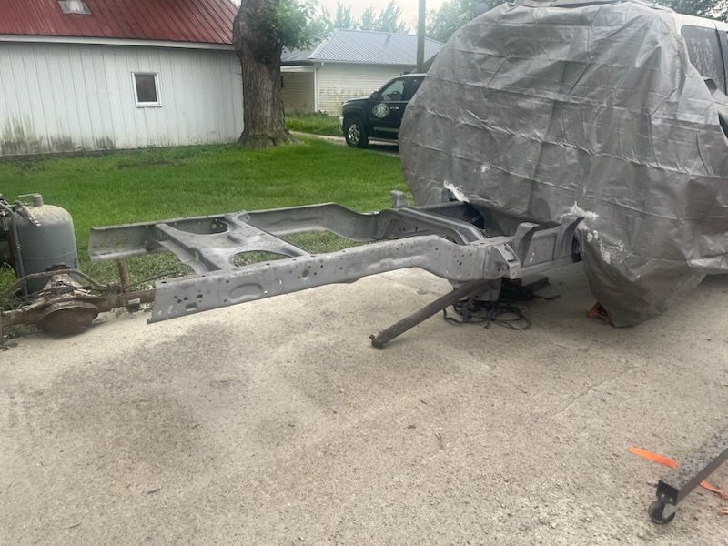 A car frame sits on a driveway, with a covered car body in the background. An axle and propane tank are nearby.