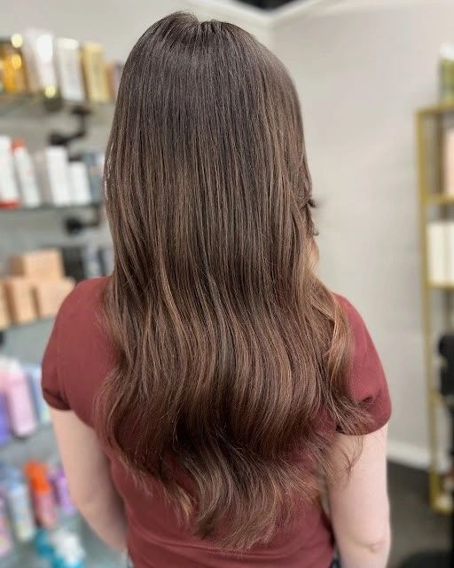 Woman with long, wavy brown hair. Highlights and lowlights. Maroon shirt. Salon background with product shelves.
