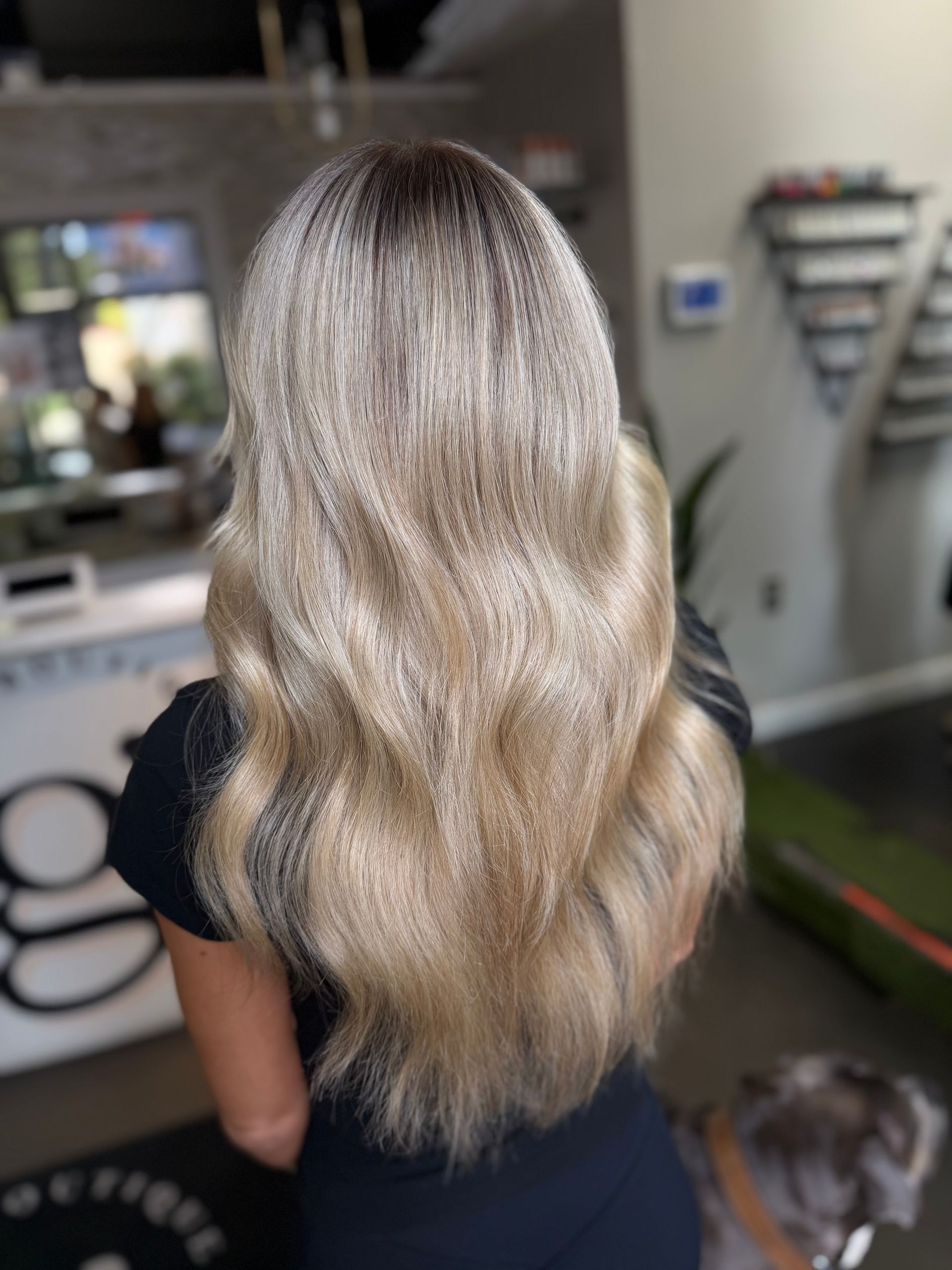 Long wavy blonde hair viewed from behind, indoors, with a small dog in the background.